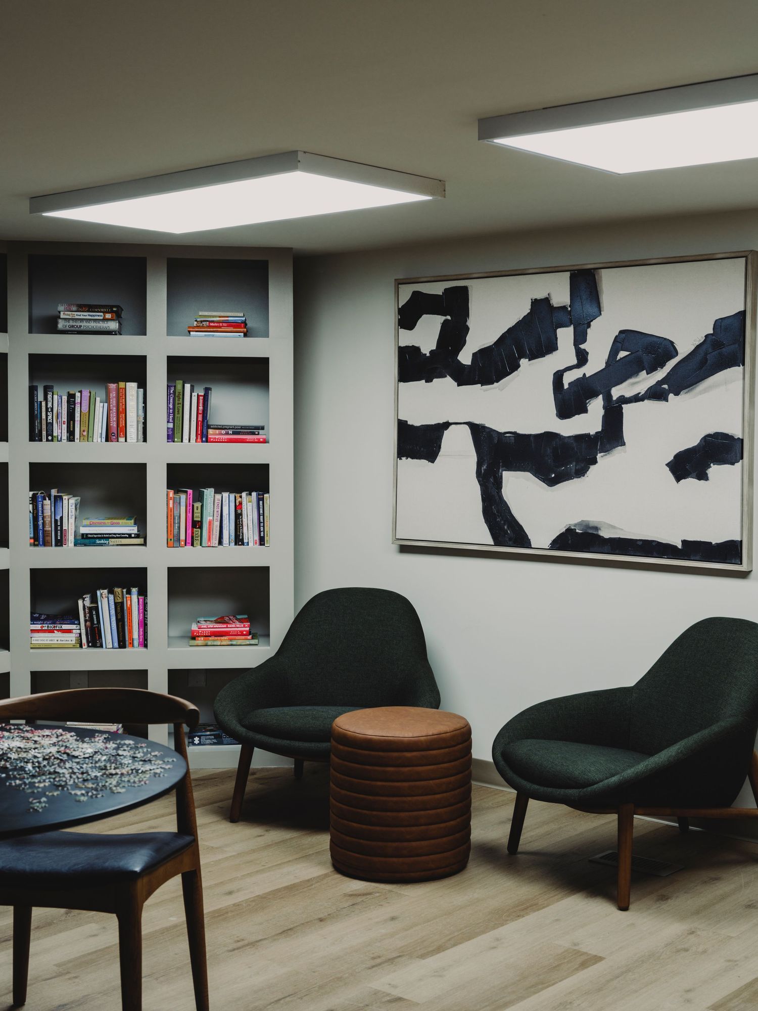Stylish lounge area with built-in bookshelf, dark green chairs, abstract wall art, and panel lighting.