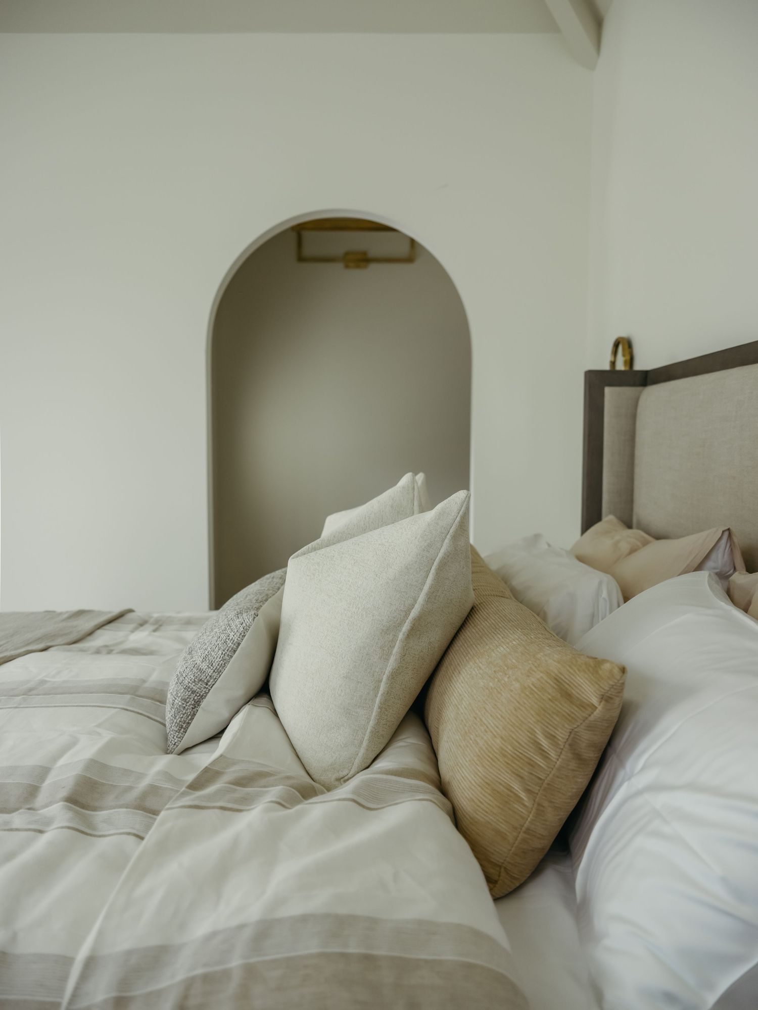Neutral bedroom with linen pillows, striped duvet, upholstered headboard, and arched wall niche with gold sconce.