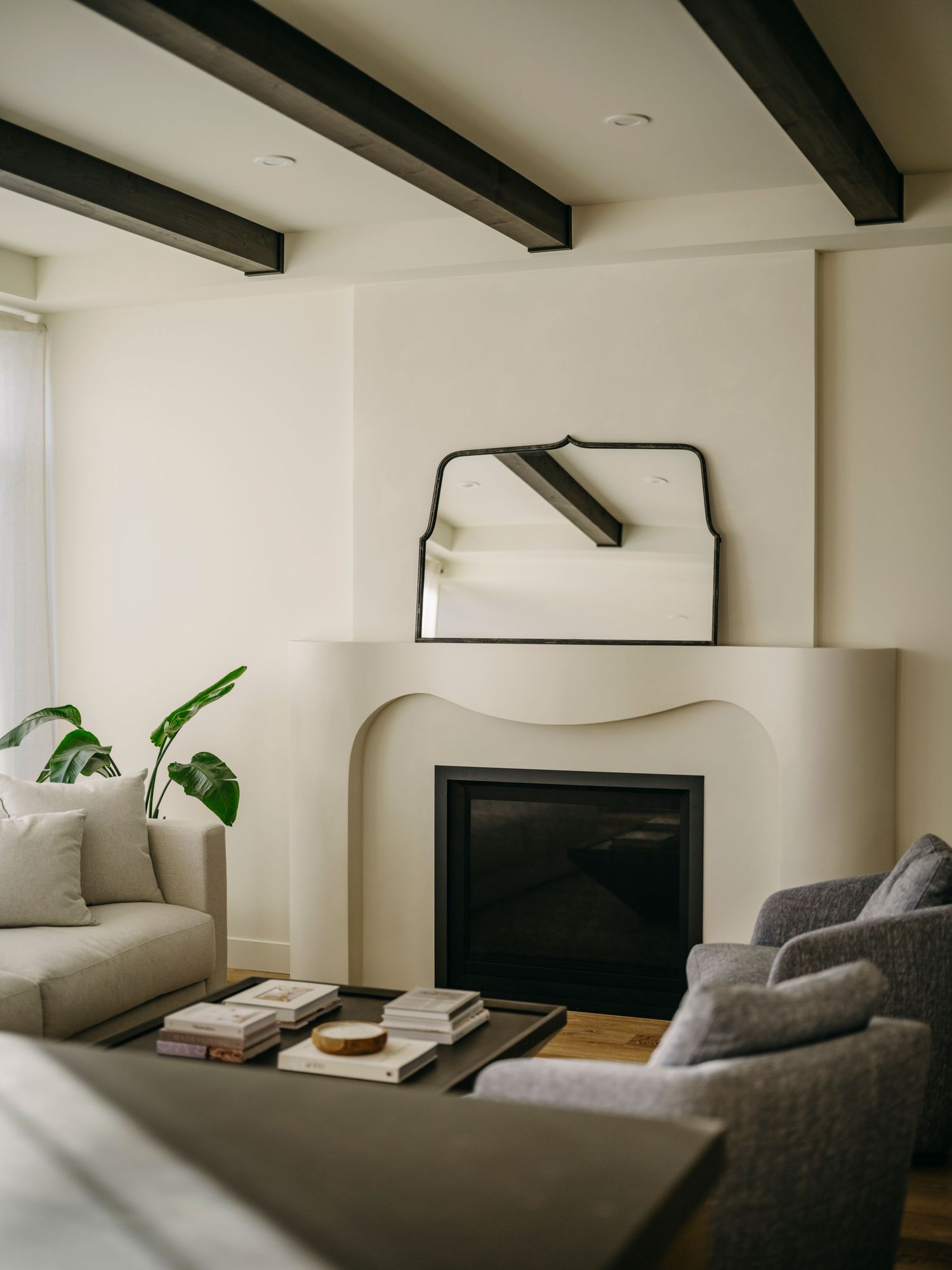 Modern living room with sculptural white fireplace, arched mirror, exposed dark wood beams, and neutral gray sofas.