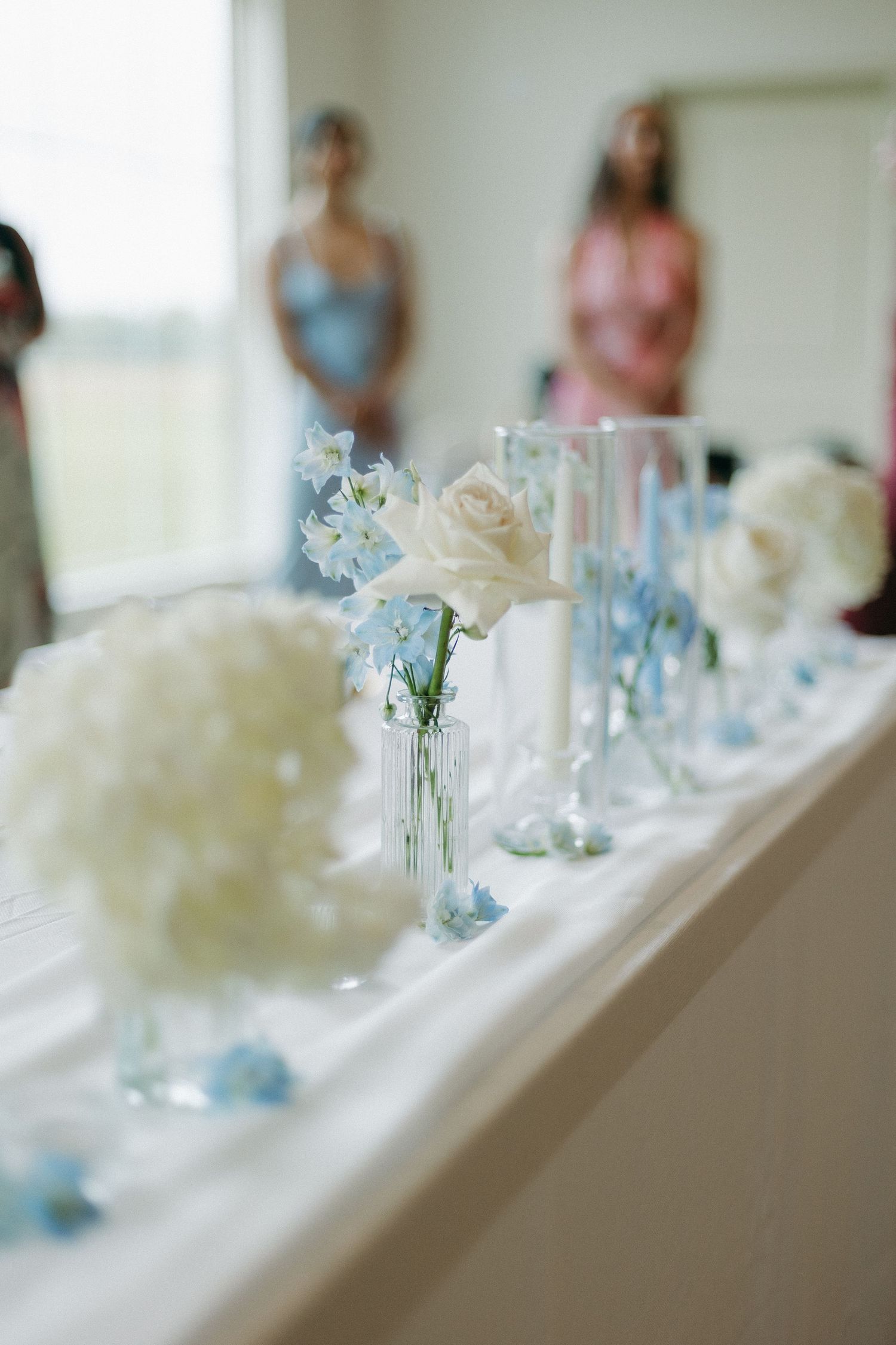 Elegant wedding table centerpiece with white roses and blue flowers in glass bud vases.