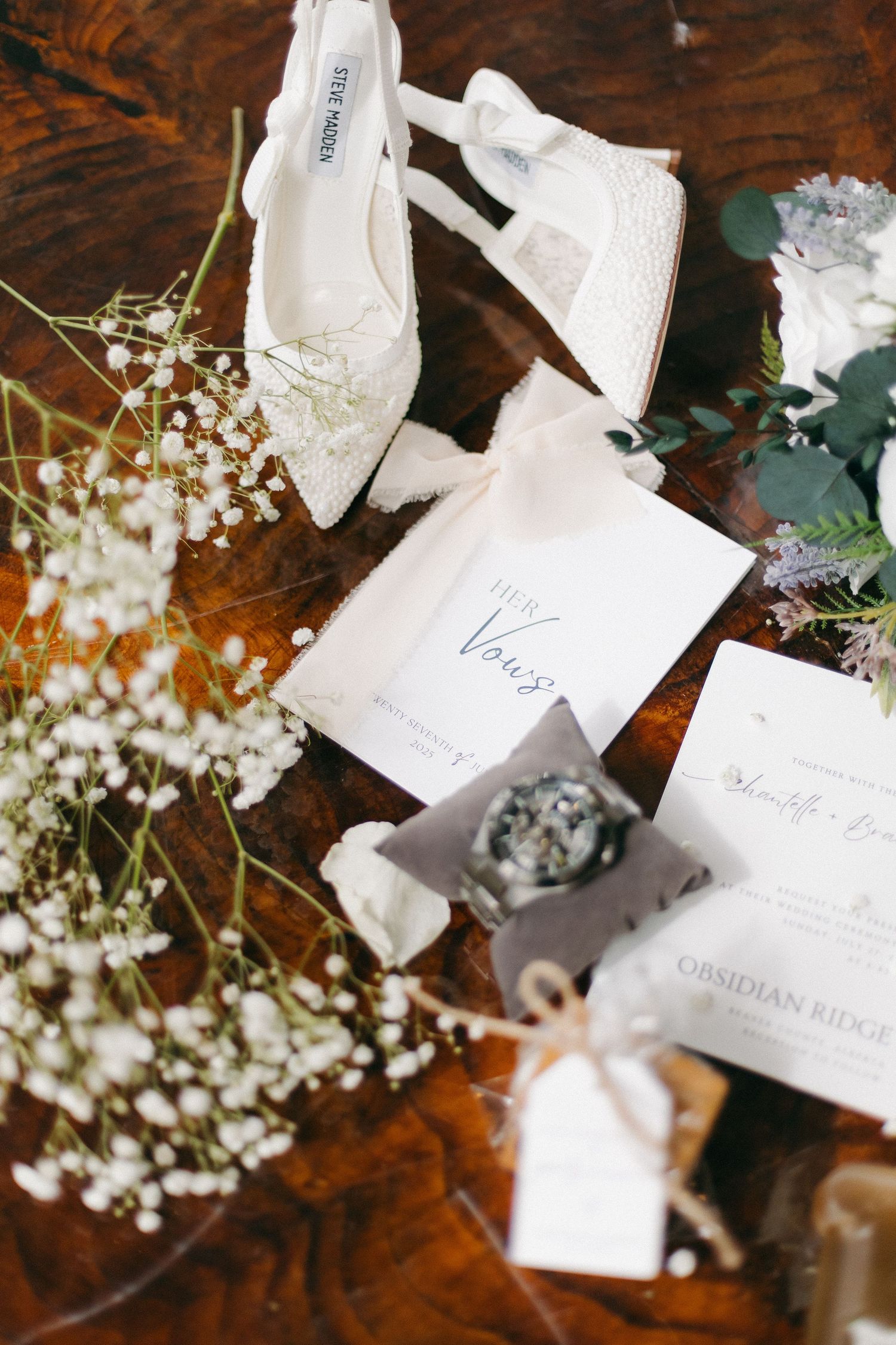 Flat lay of white wedding heels, vow booklets, ring box, baby's breath flowers, and stationery on wooden surface.
