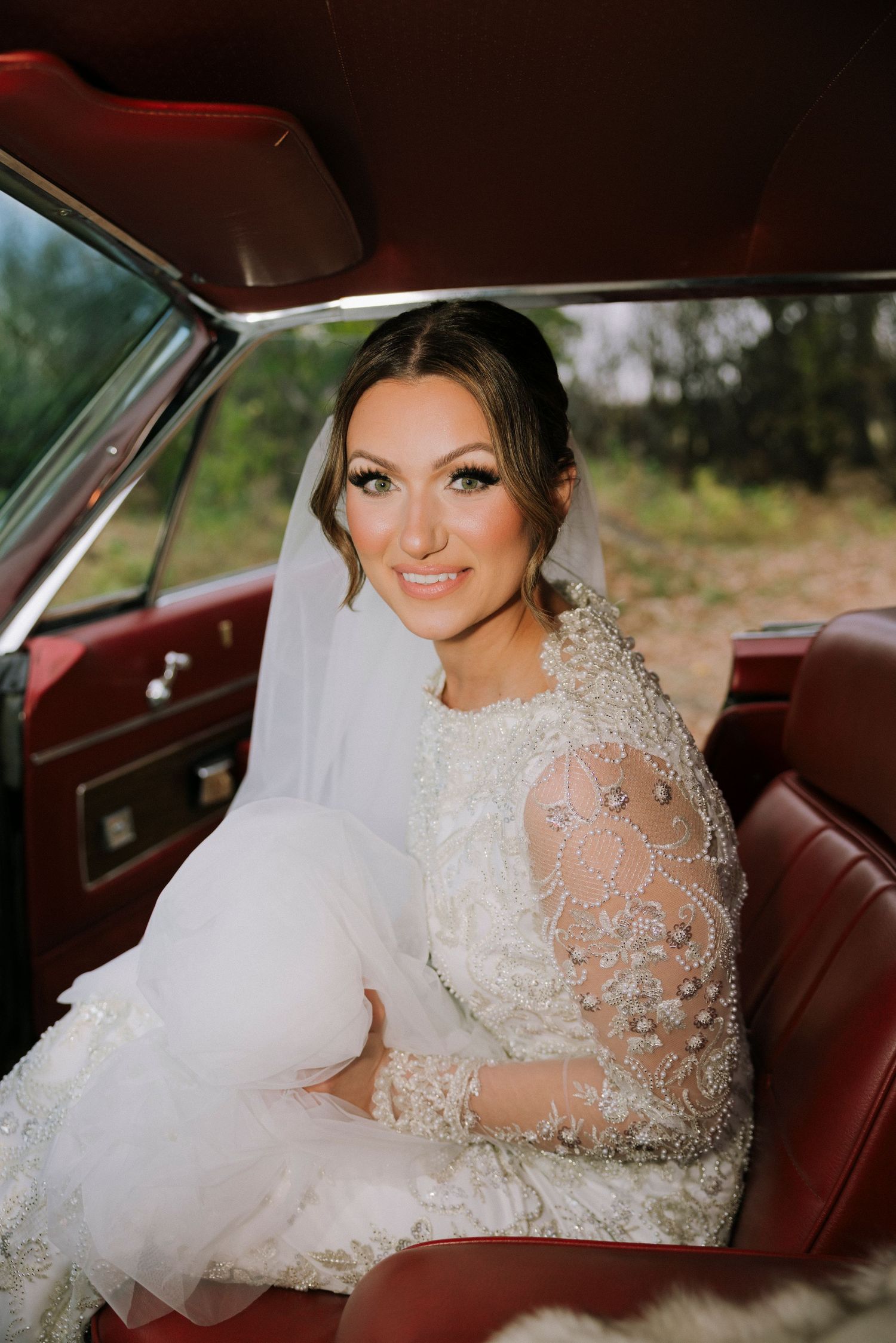 Radiant bride in beaded lace wedding dress sitting in vintage red leather car interior.