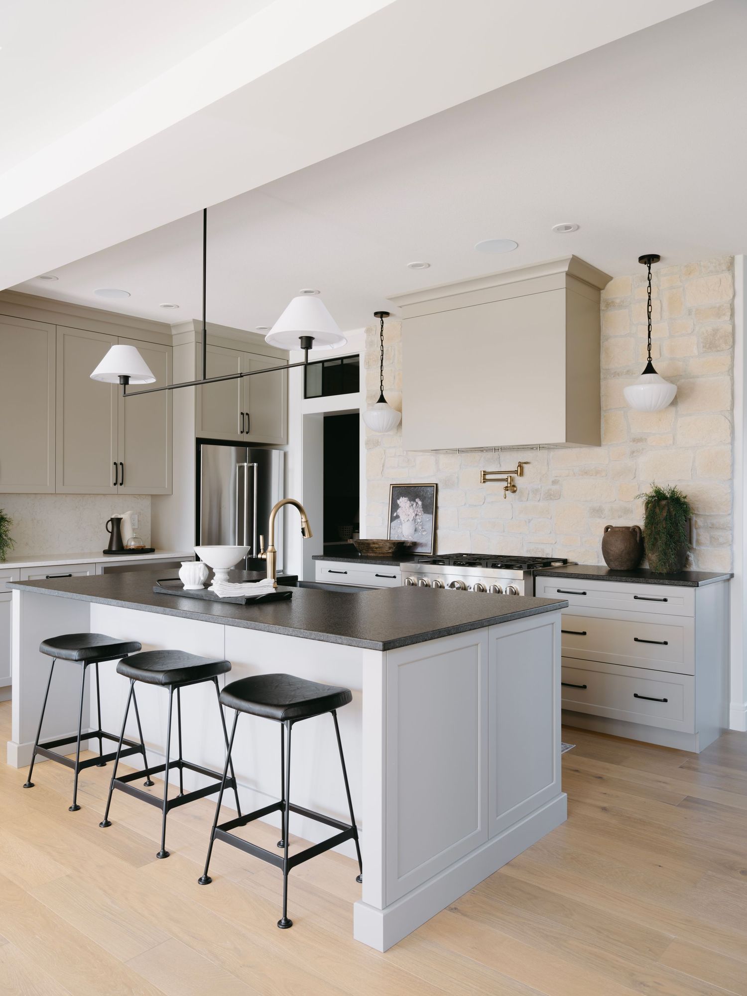Modern kitchen with grey shaker cabinets, dark countertops, black bar stools, pendant lights, and stone backsplash.