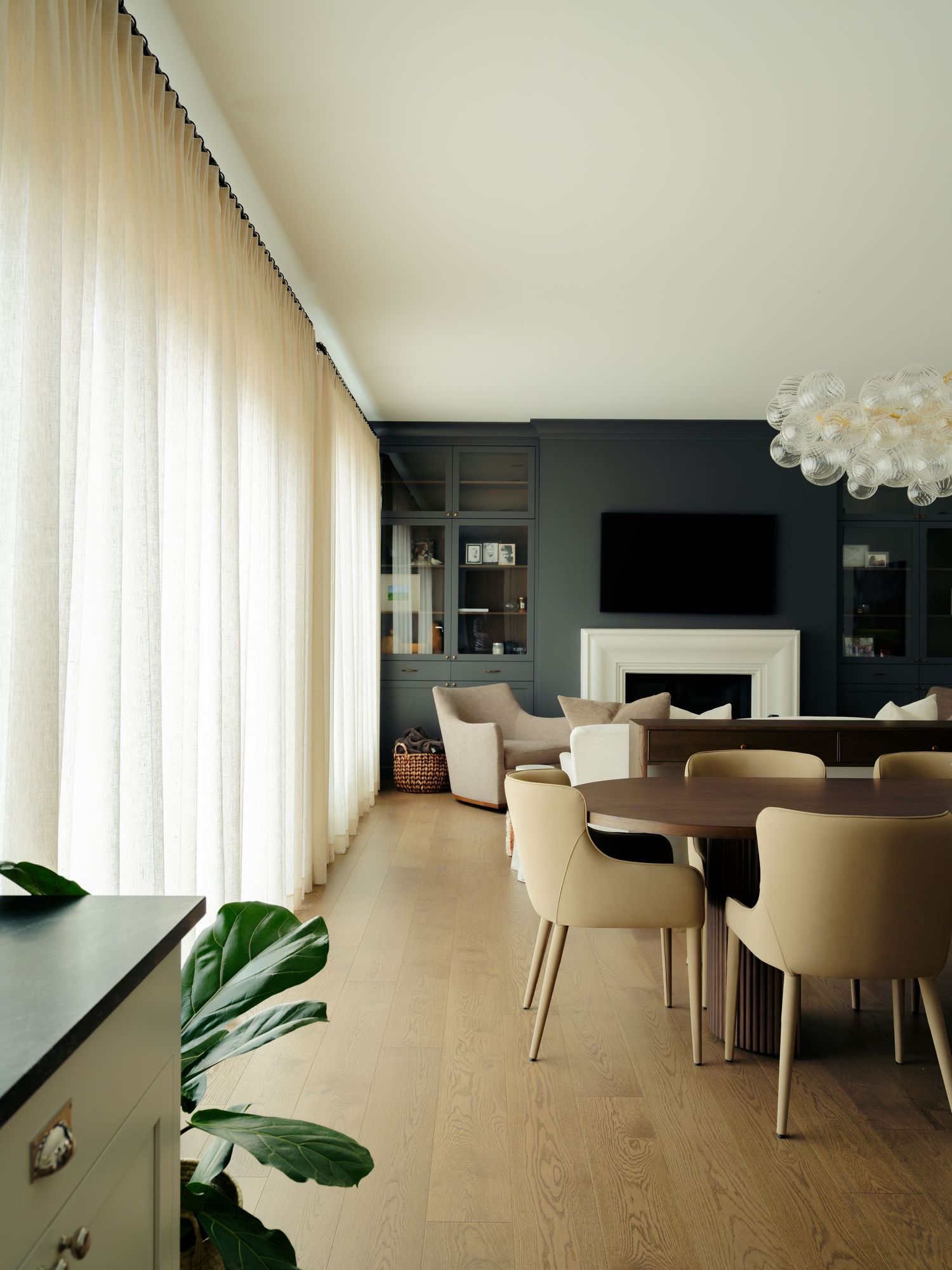 Elegant open-plan living and dining room with dark walls, fireplace, beige chairs, sheer curtains, and light wood floors.