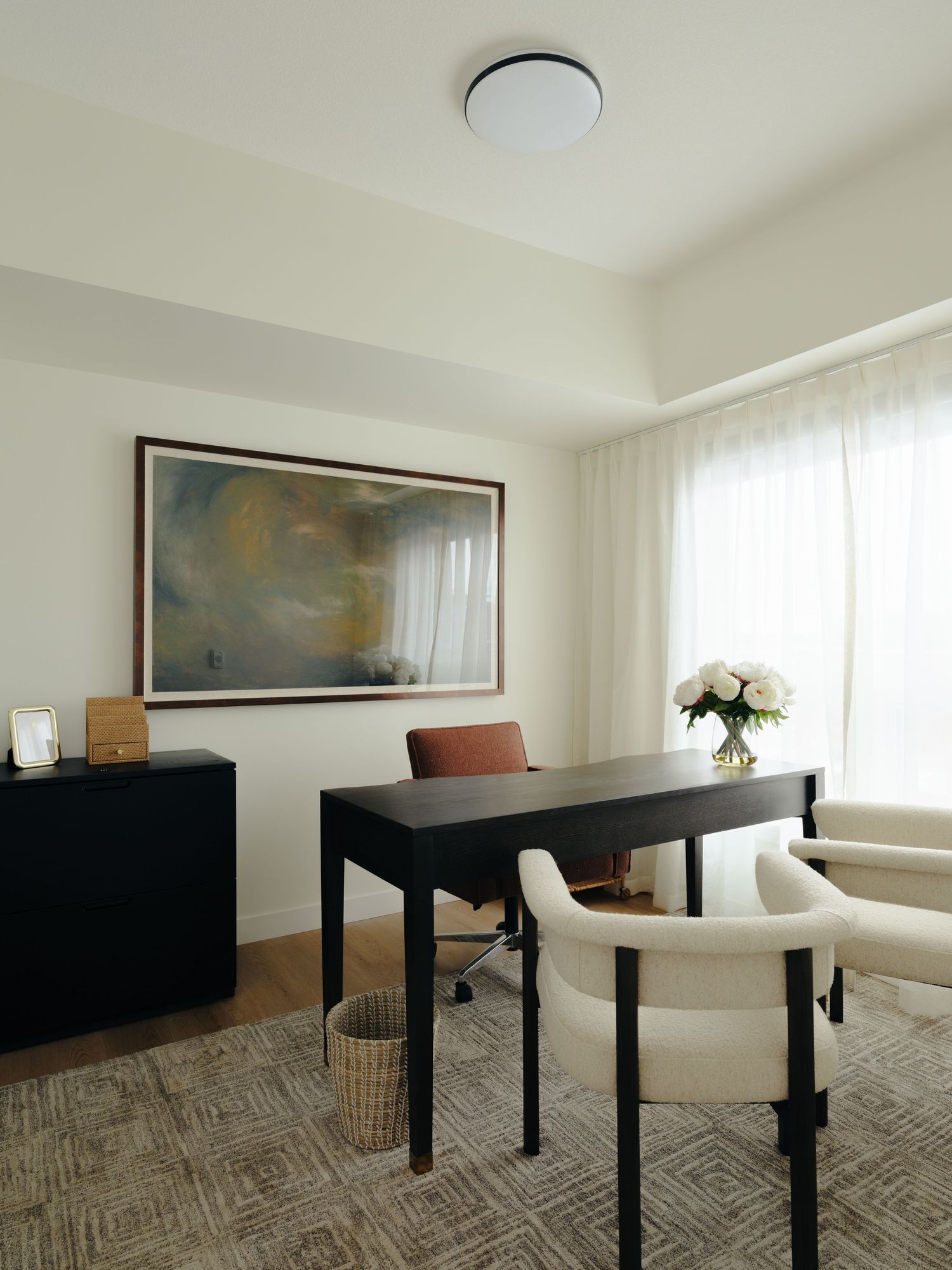 Minimalist home office with black desk, cream accent chairs, large landscape painting, and sheer white curtains.