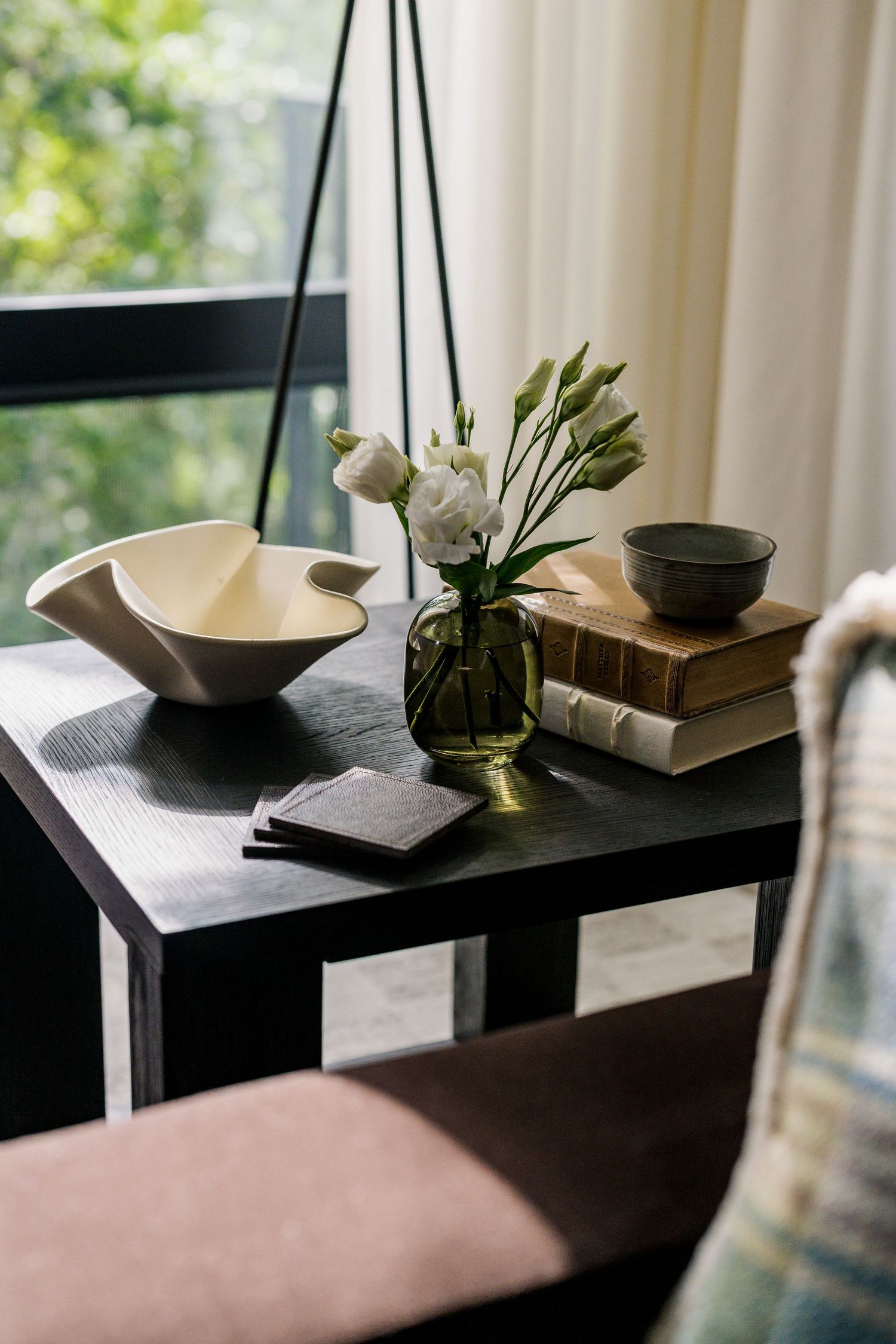 Dark wood side table styled with white flowers in green vase, stacked books, ceramic bowl, and decorative dish near window.