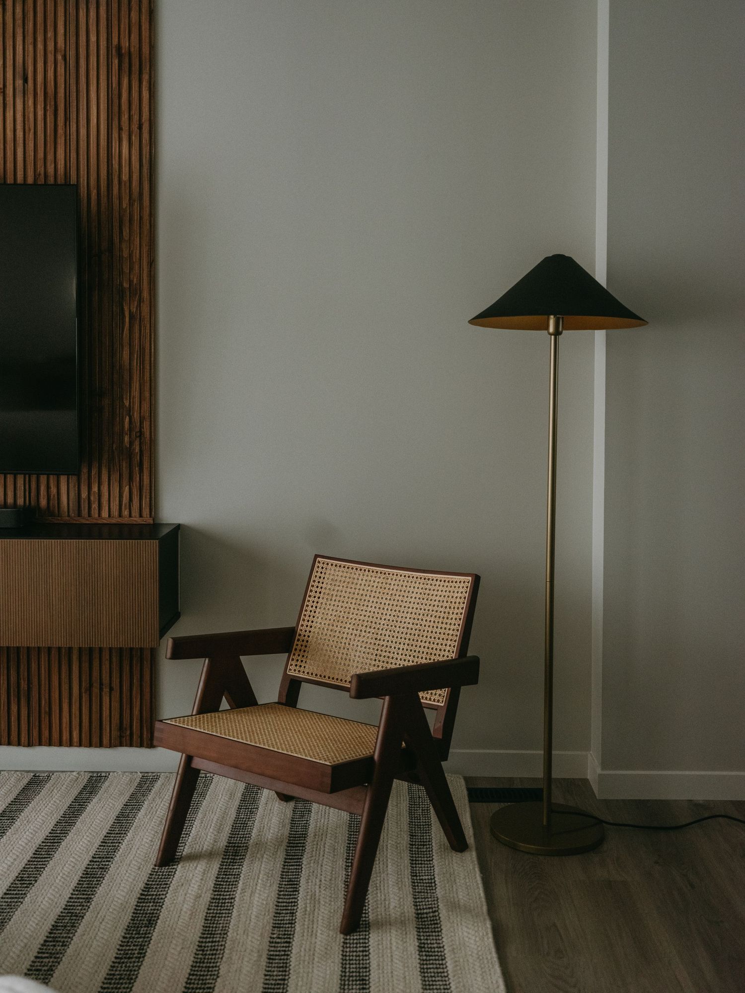Cane and walnut lounge chair beside a brass floor lamp with dark shade on a striped rug.
