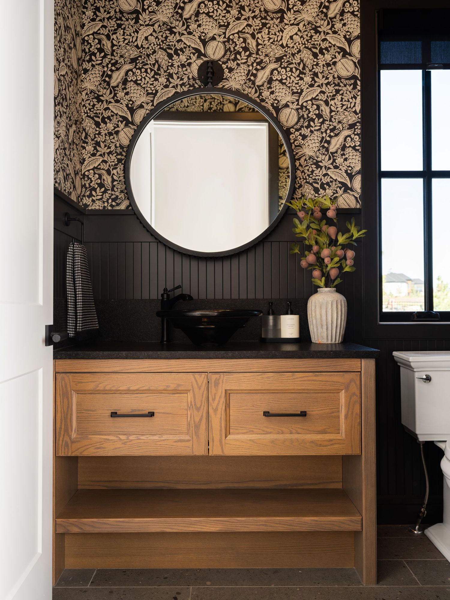 Oak bathroom vanity with black countertop, round mirror, vessel sink, and bold floral wallpaper accent wall.