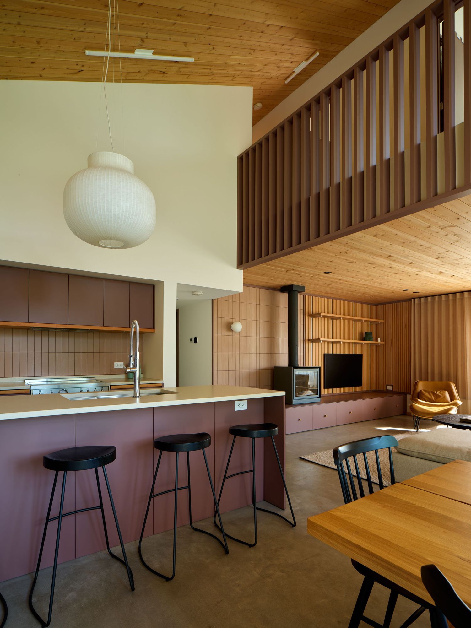 Open-plan interior with pink kitchen island, black stools, wood ceiling, loft railing, and freestanding stove.