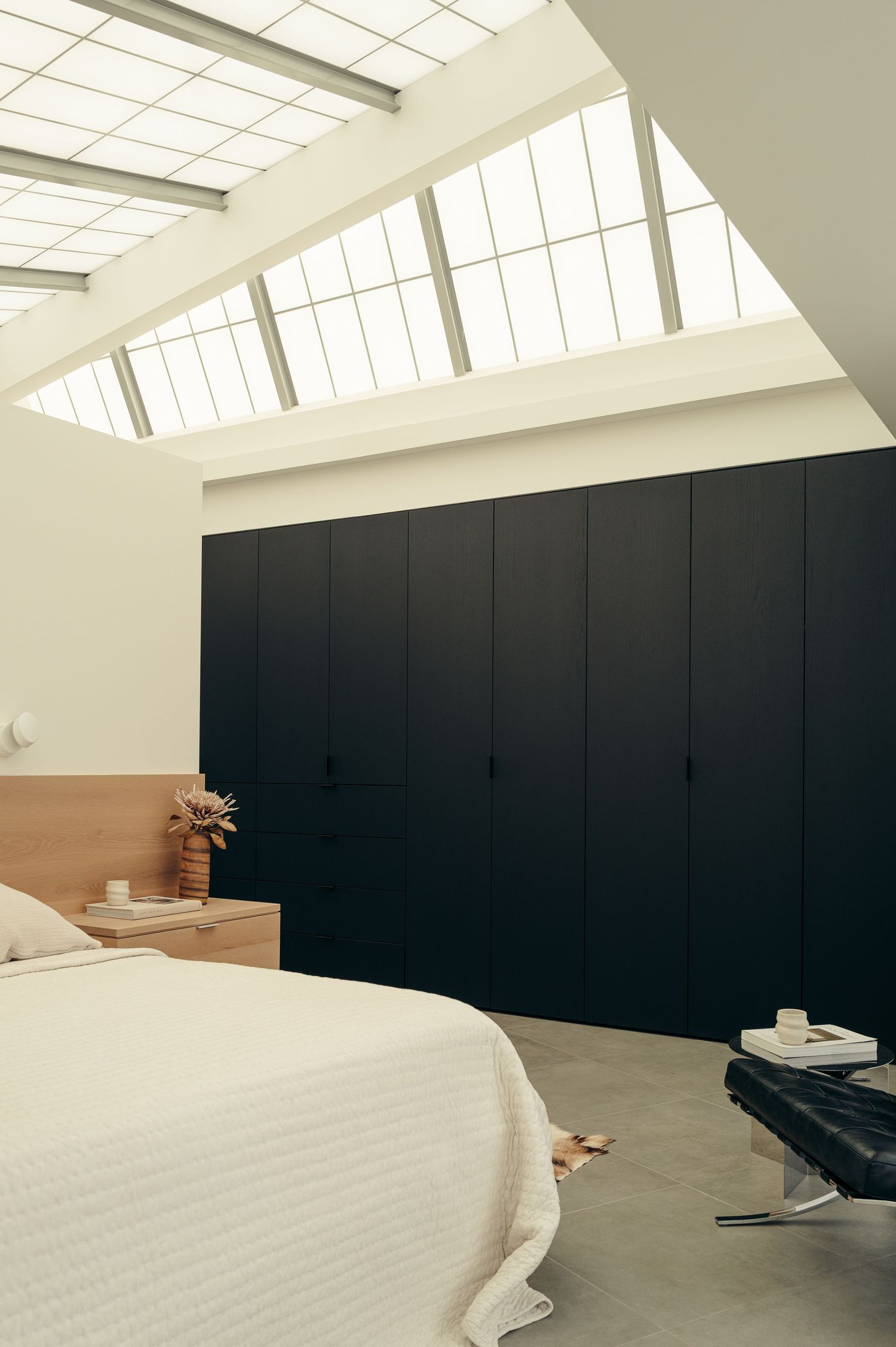Minimalist bedroom with large skylights, floor-to-ceiling black wardrobe, white bedding, and concrete floors.