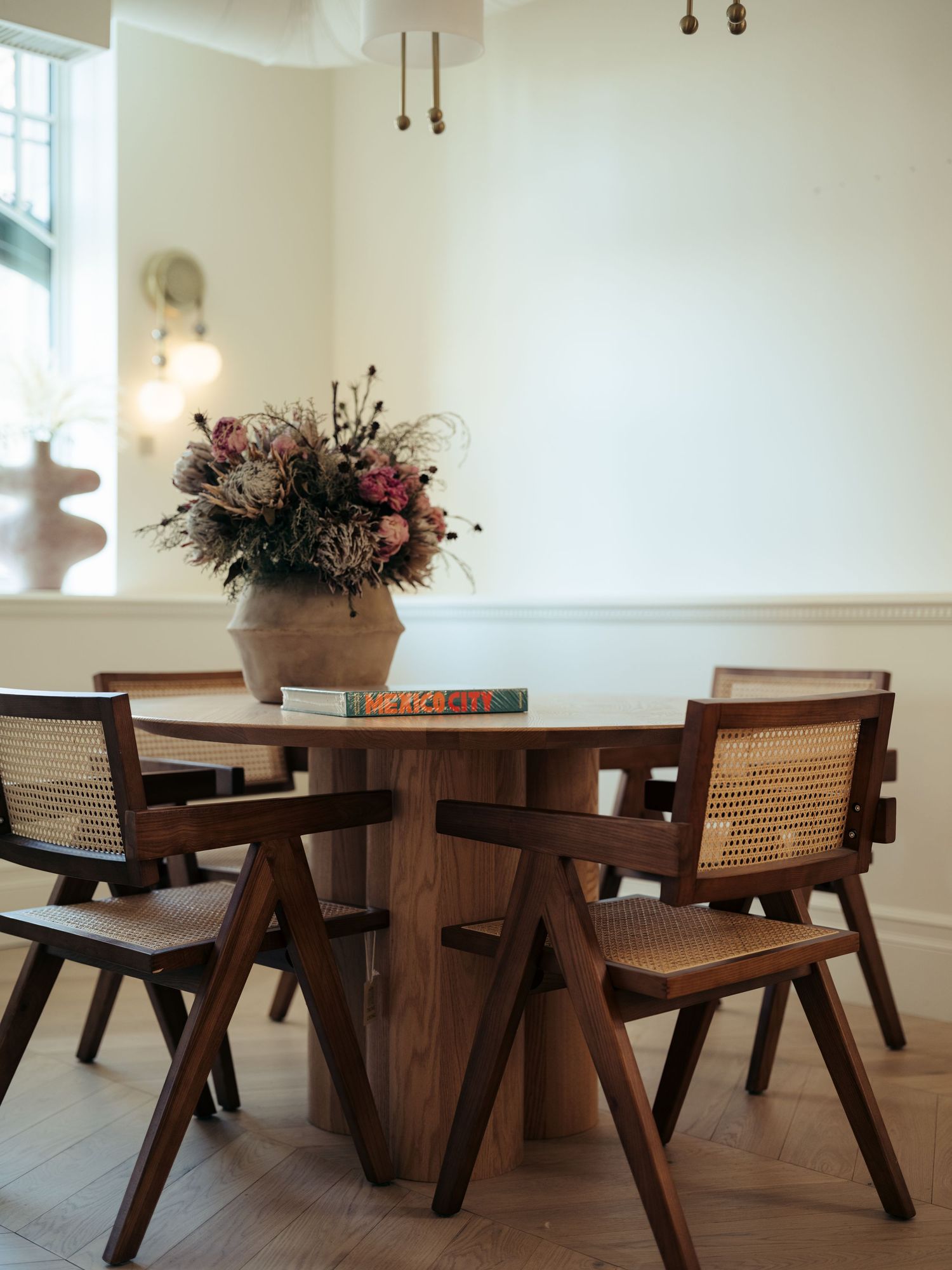 Rustic dining area with round wood table, cane-back chairs, herringbone floor, and floral centerpiece arrangement.