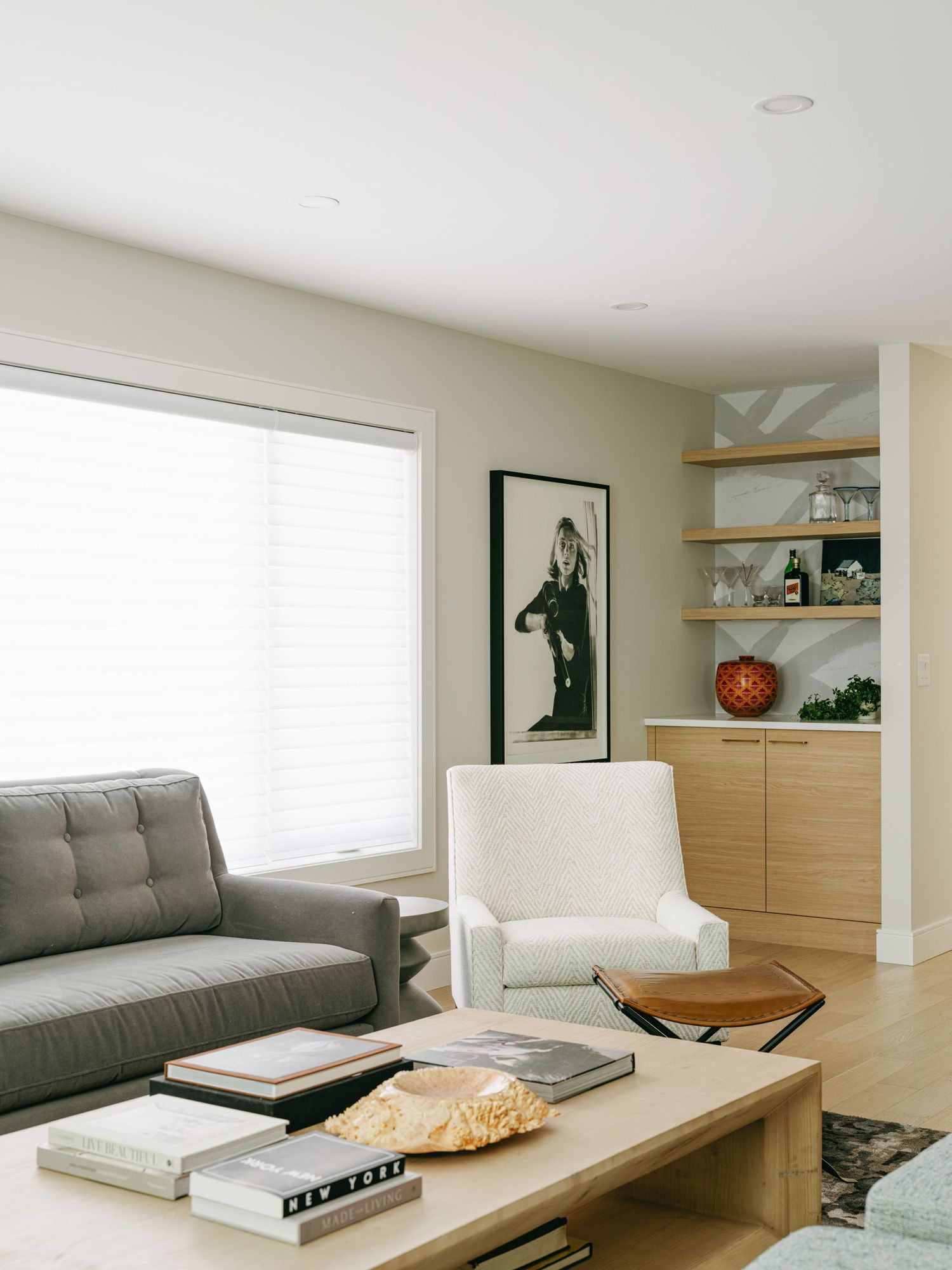 Minimalist living room with gray tufted sofa, light wood coffee table, built-in shelving, and large window.