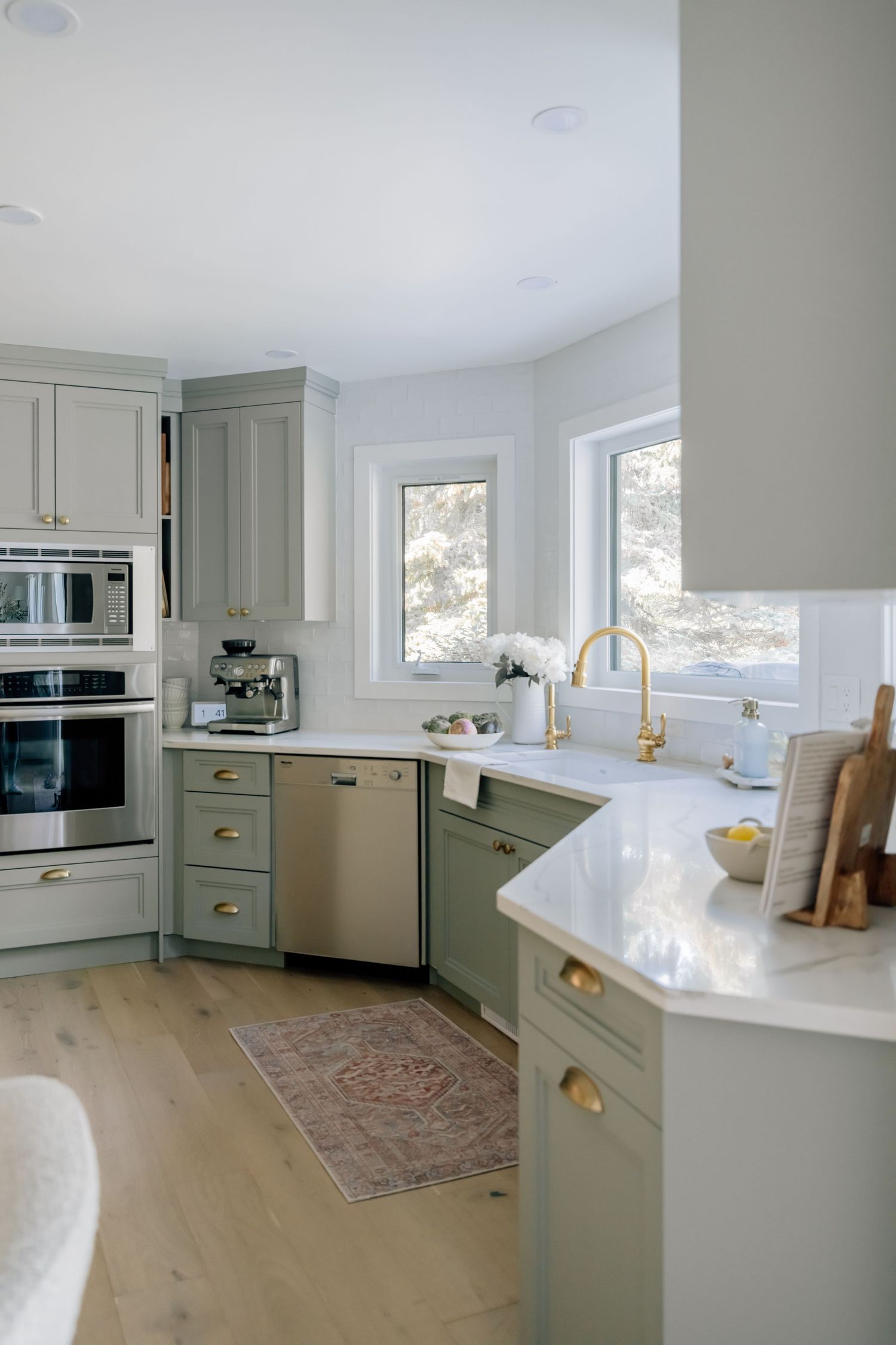 Sage green kitchen cabinets with brass hardware, white quartz countertops, stainless steel appliances, and light hardwood floors.