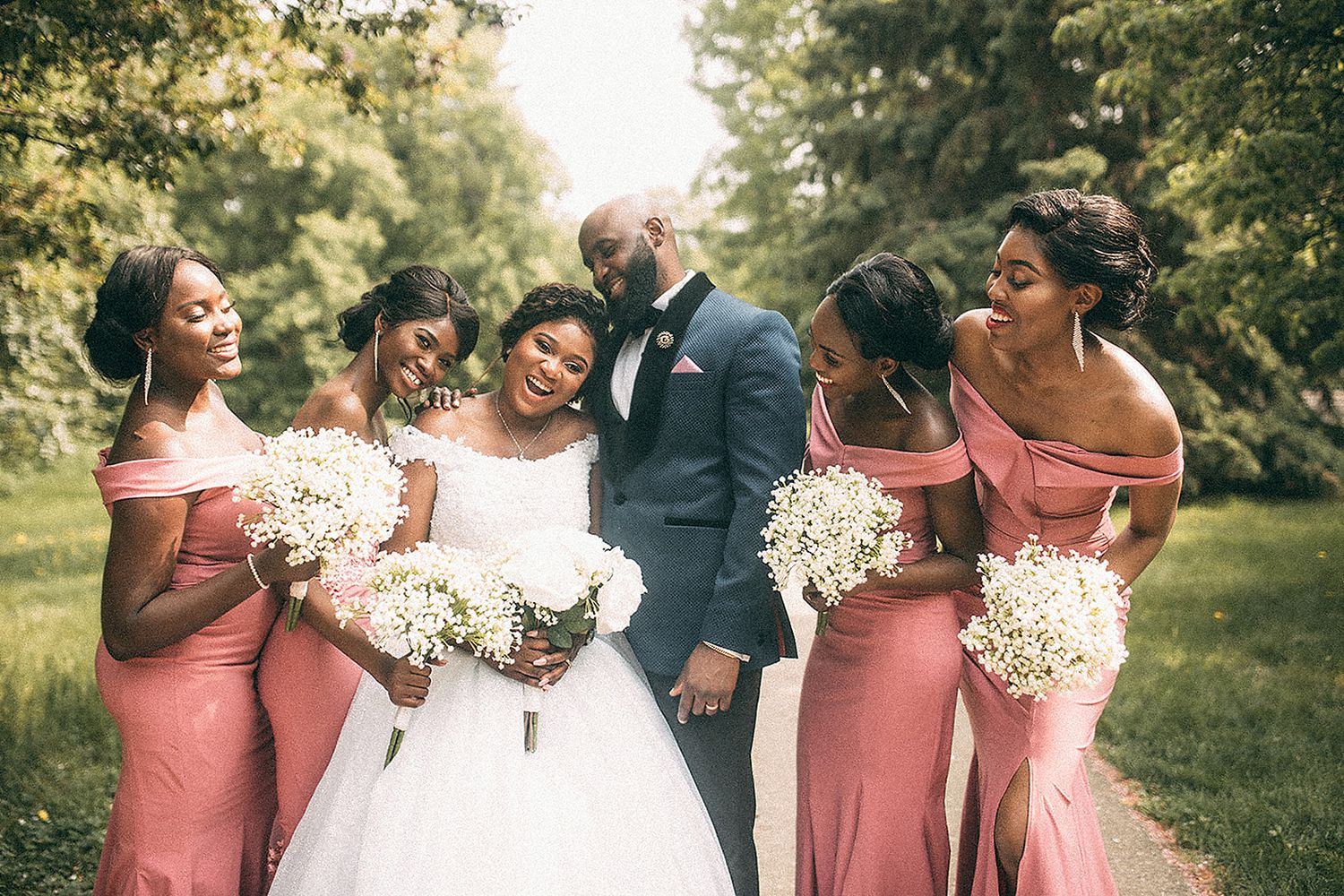 Bride in white gown with groom and four bridesmaids in pink off-shoulder dresses holding bouquets outdoors.
