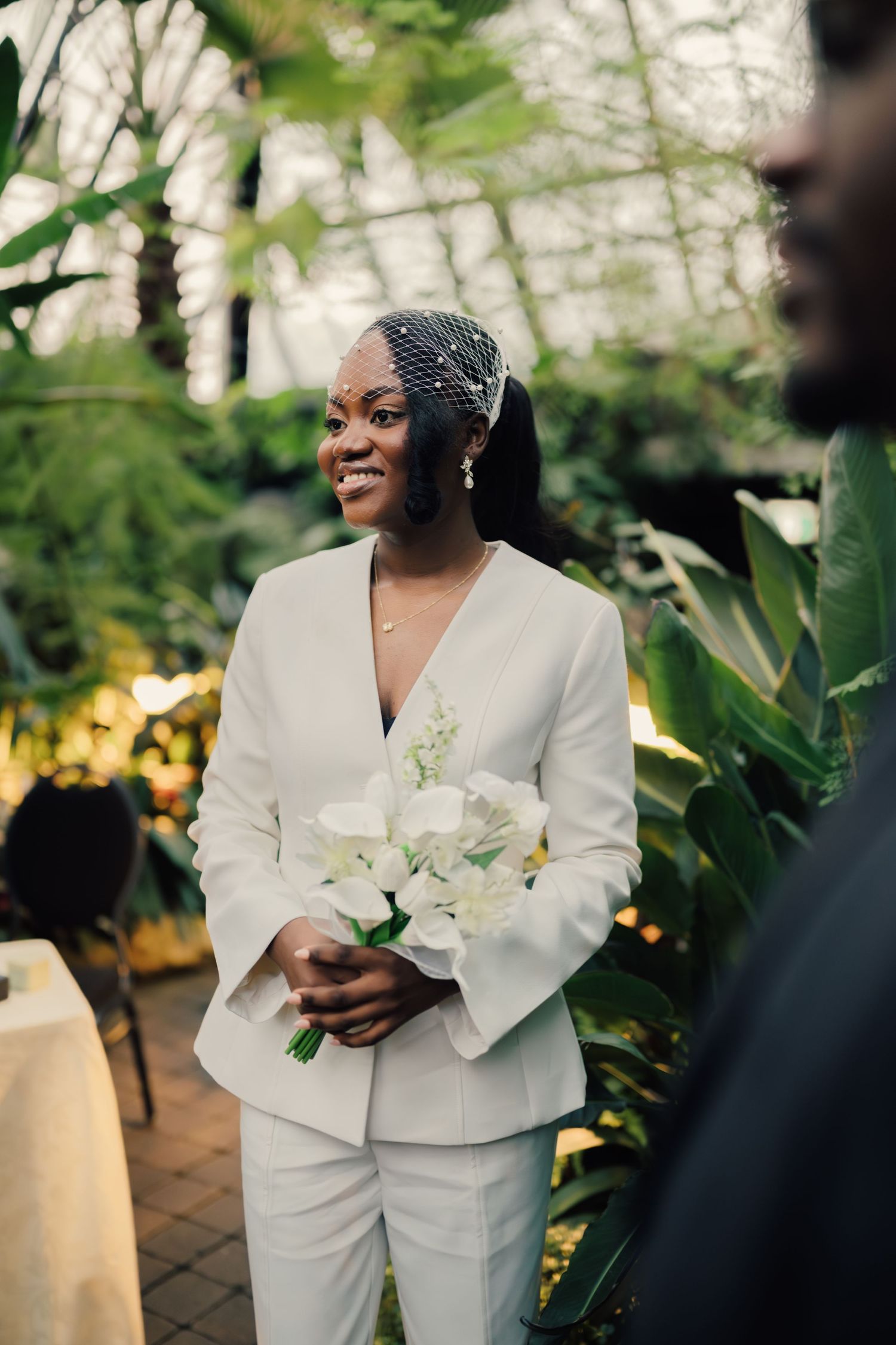 Radiant bride in white pantsuit and birdcage veil holding white bouquet in tropical garden setting.
