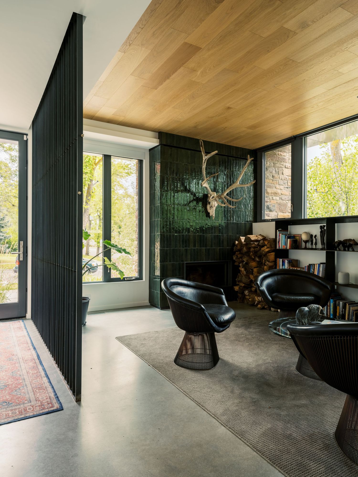 Modern living room with green tile fireplace, black leather chairs, wood ceiling, and concrete floor.