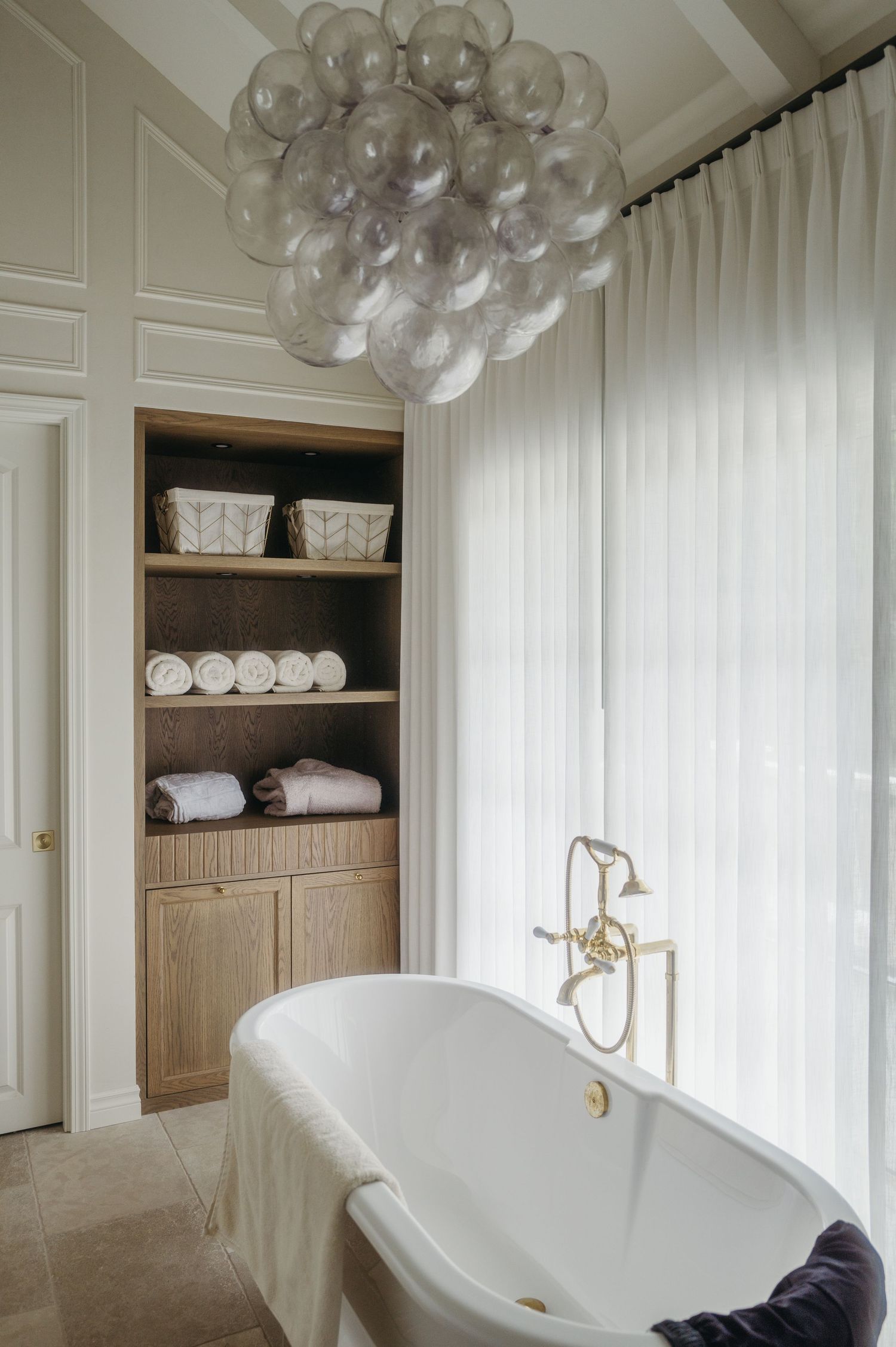 Elegant bathroom with freestanding white soaking tub, brass fixtures, bubble chandelier, sheer curtains, and built-in wooden shelving.