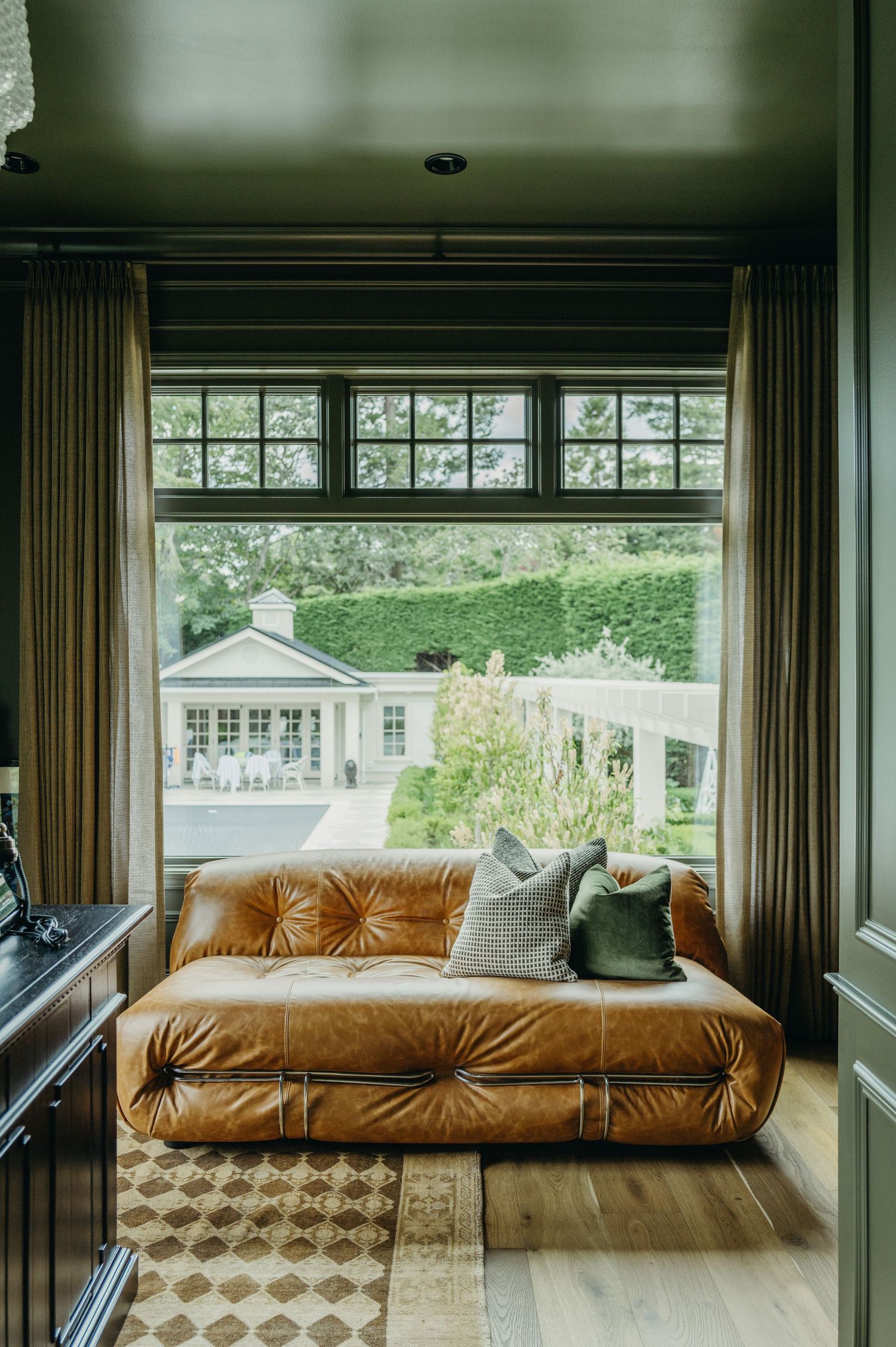 Tufted cognac leather sofa by large windows overlooking lush garden, with checkered rug and neutral cushions.