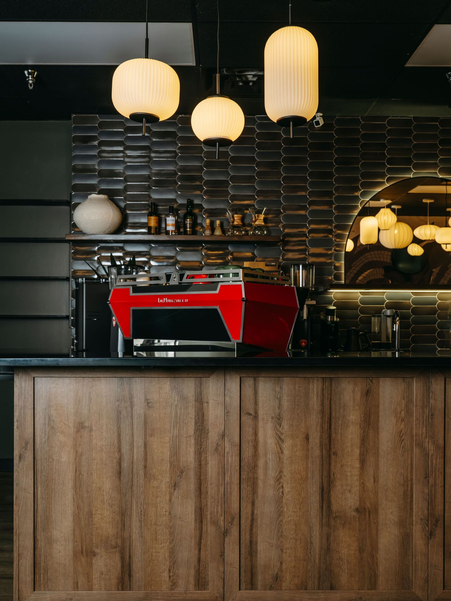 Red La Marzocco espresso machine on wood counter with globe pendant lights and dark hexagonal tile backsplash.