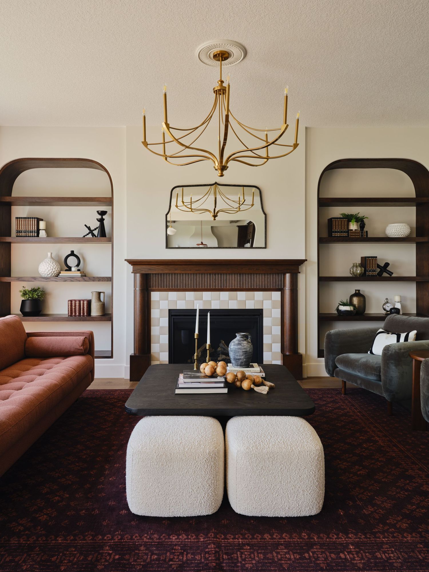 Elegant living room with gold chandelier, arched bookshelves, tiled fireplace, leather sofas, and white boucle ottomans on a dark rug.