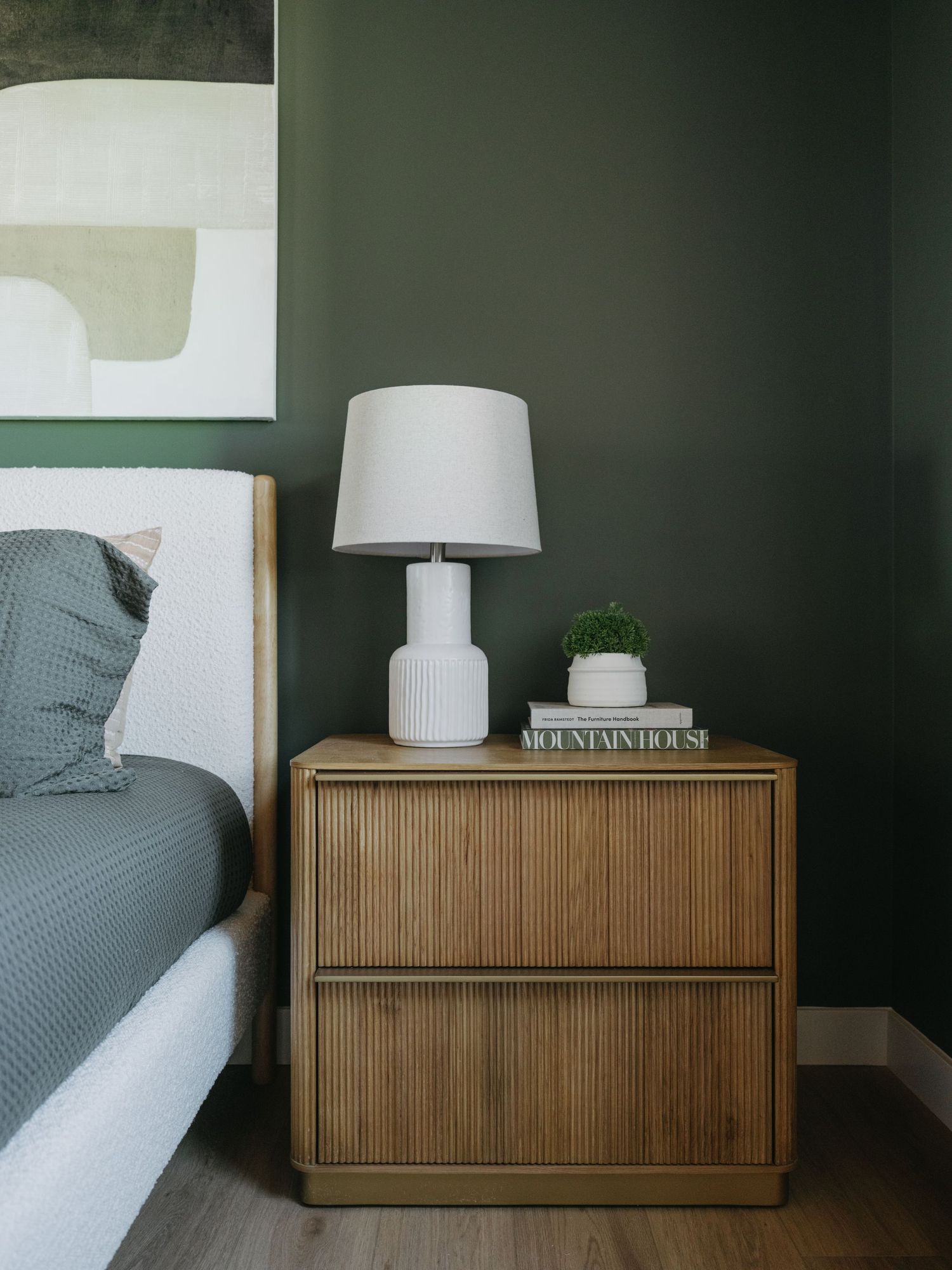 Oak nightstand with white ribbed lamp, small plant, and book against dark green bedroom wall.