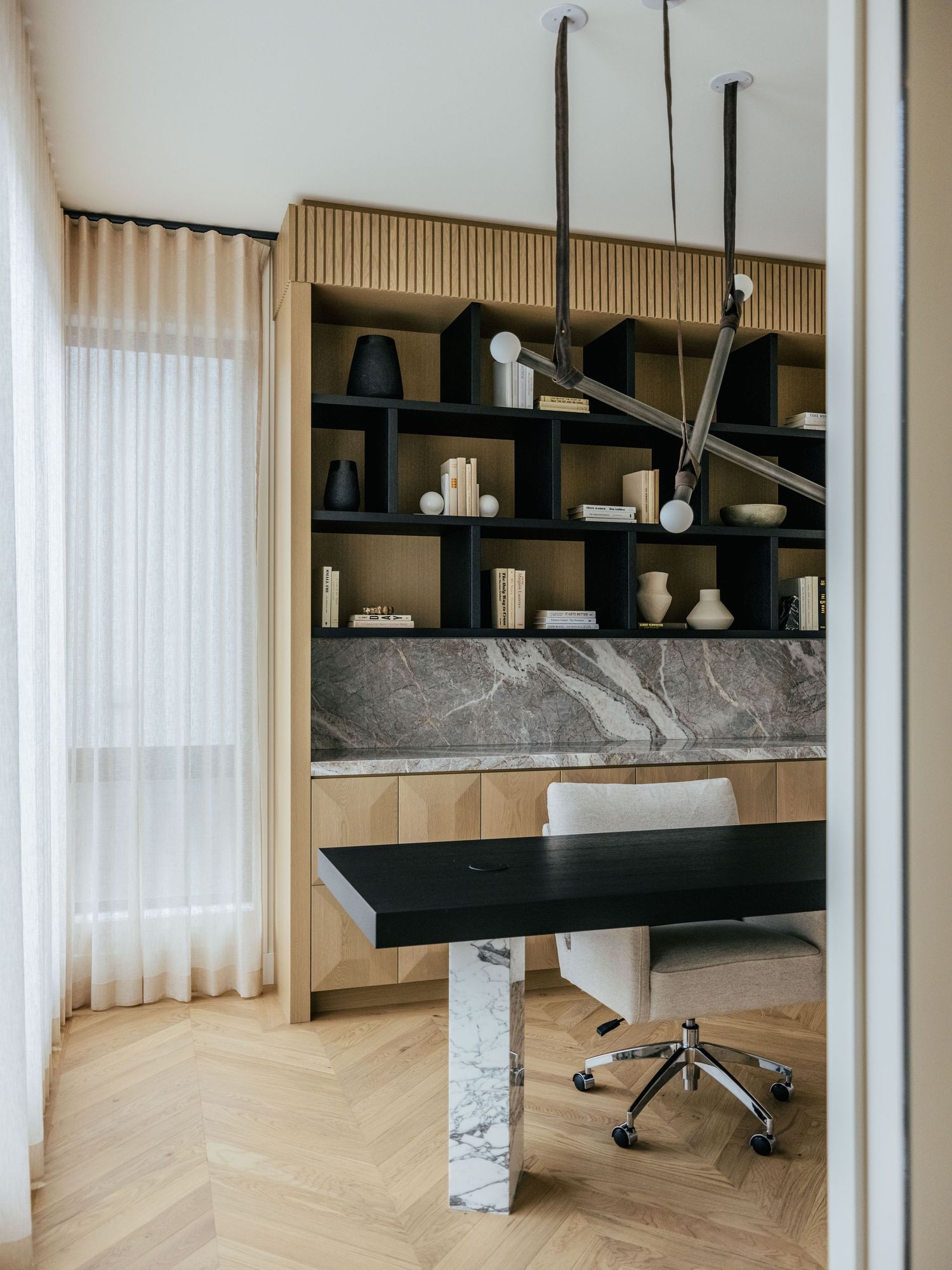 Modern home office with marble desk, black shelving unit, herringbone wood floors, and sculptural pendant light.