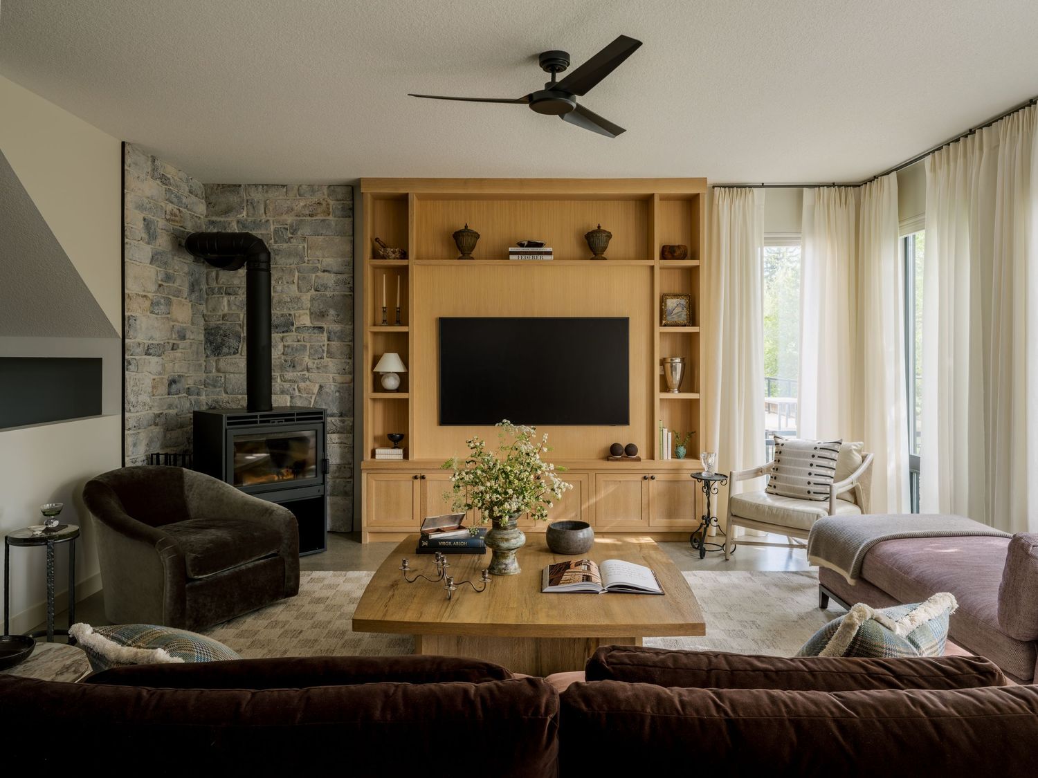 Cozy living room with wood built-ins, wall-mounted TV, stone fireplace, ceiling fan, and neutral furniture arrangement.
