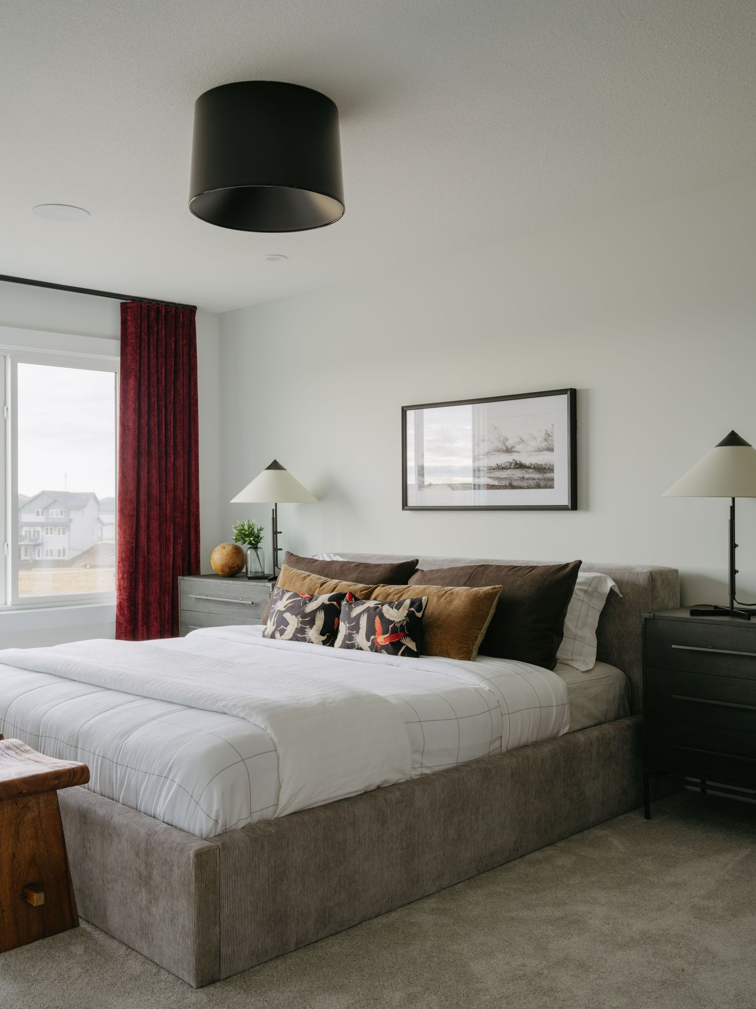 Modern bedroom with gray upholstered bed, layered pillows, black pendant light, and burgundy curtains by a window.
