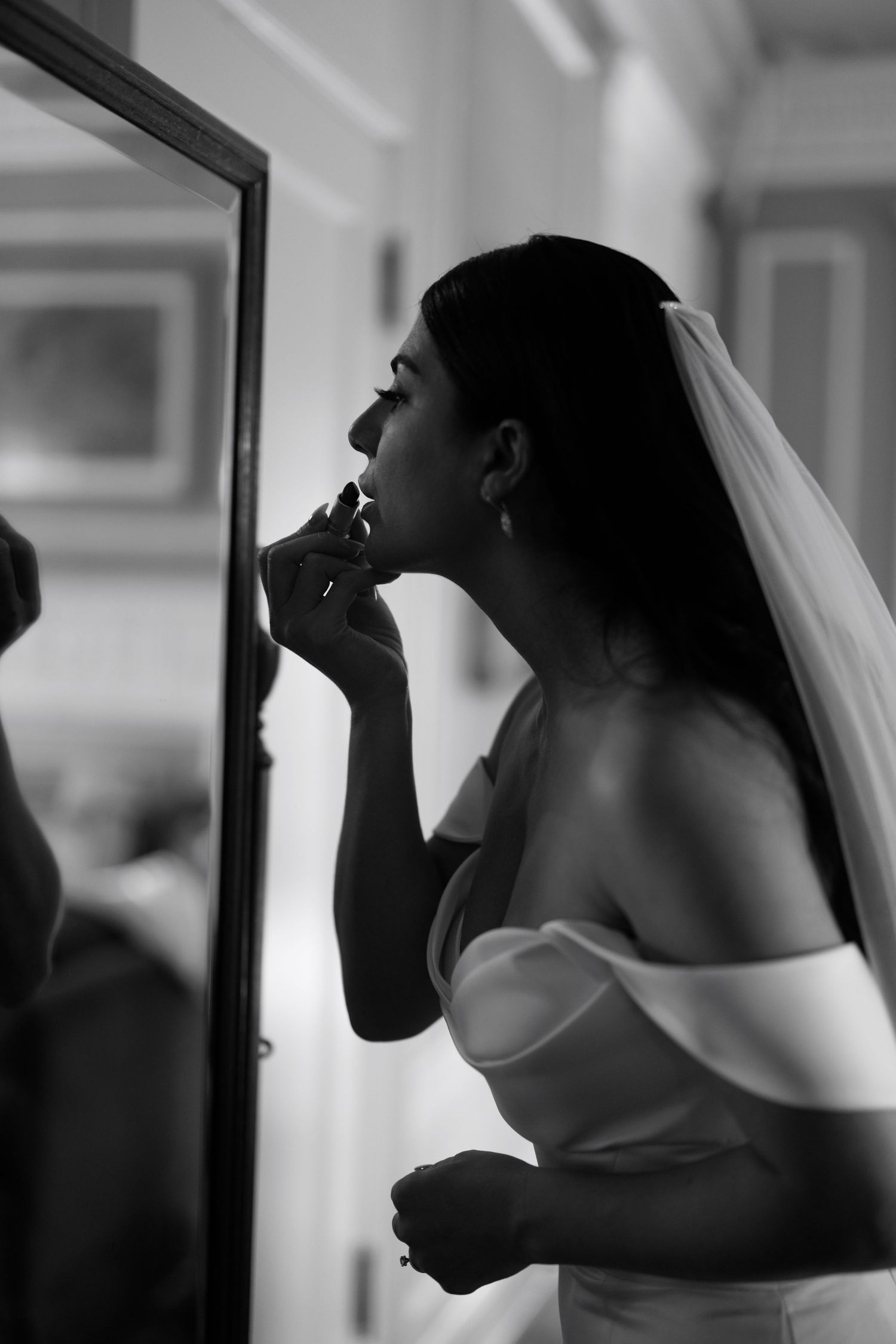 Black and white photo of bride in off-shoulder gown applying lipstick in front of mirror.