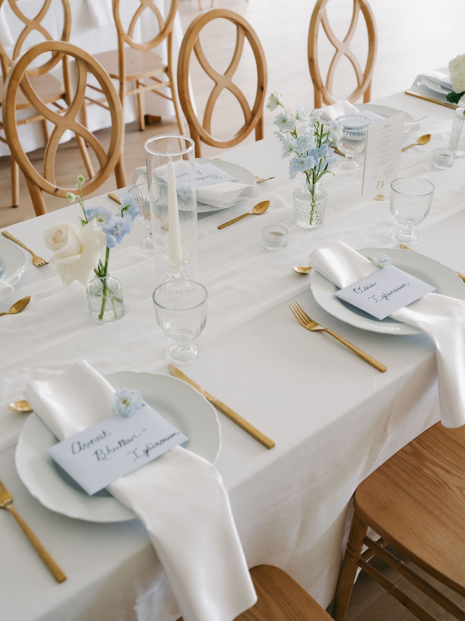Elegant wedding reception table with white linens, gold cutlery, and blue floral centerpieces.