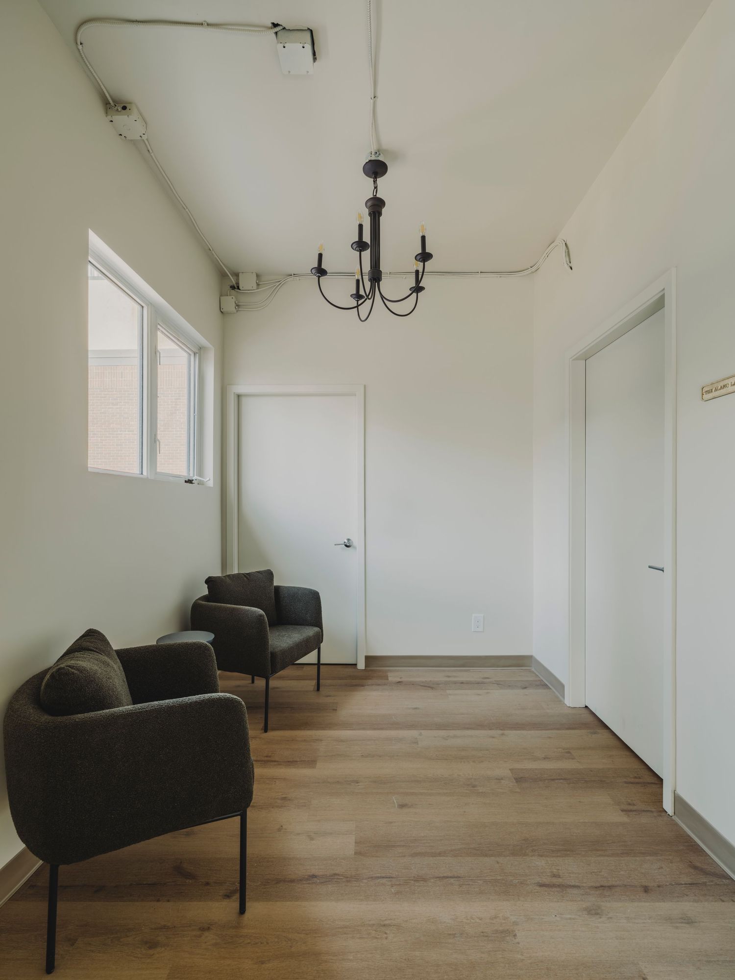 Minimalist waiting room with two dark armchairs, chandelier, white walls, and light hardwood floors.