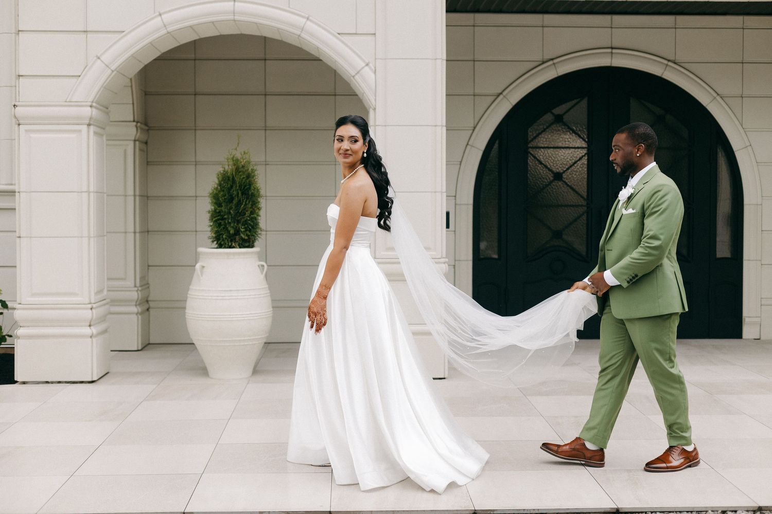 Bride in a flowing white gown looks back as groom in a sage green suit holds her veil outside a white building.
