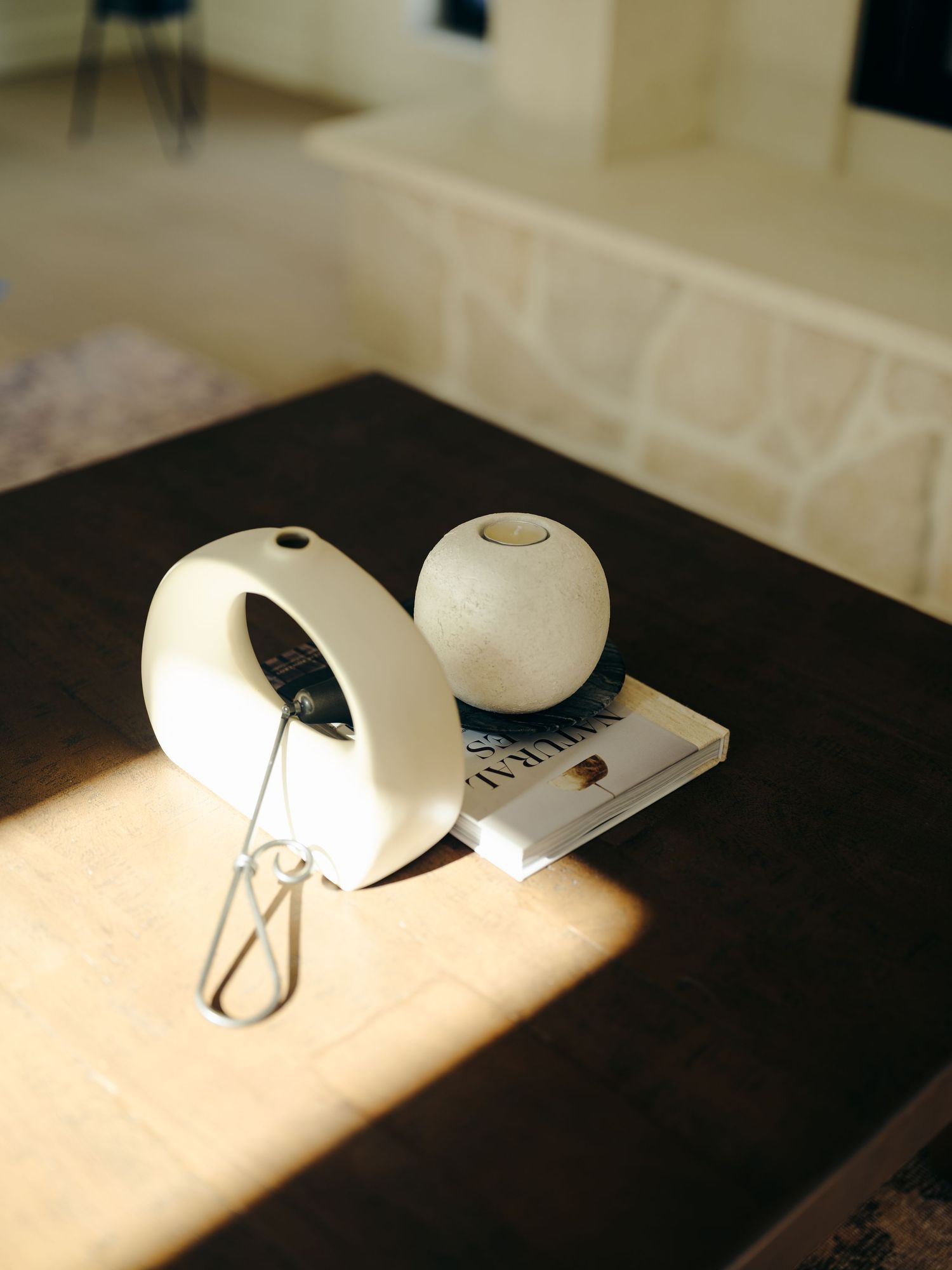 White ceramic donut sculpture and sphere vase styled on a dark wood table in sunlit interior.