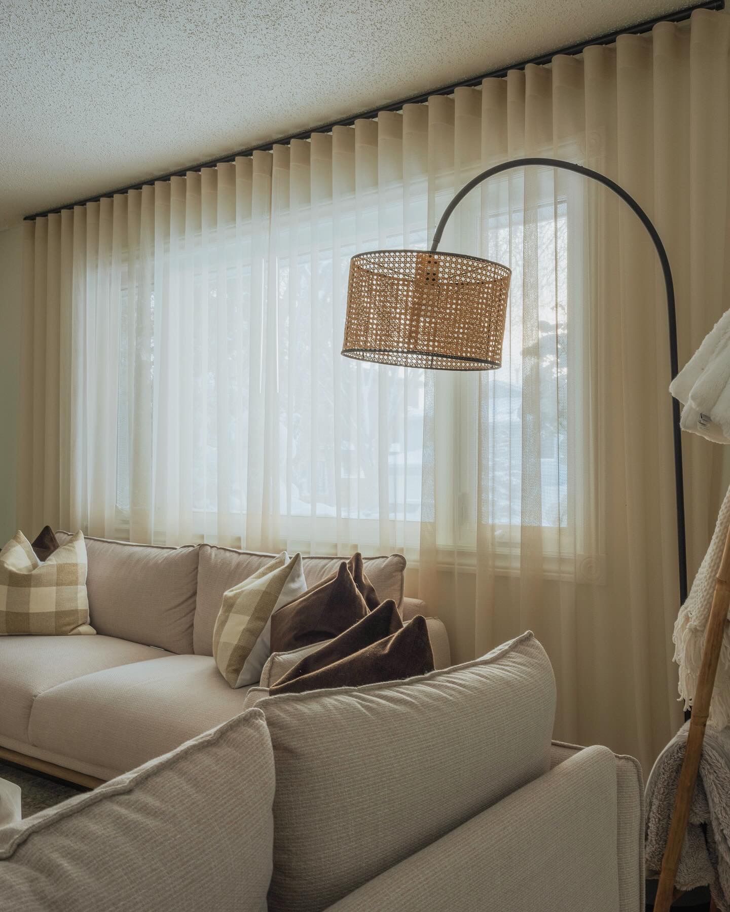 Neutral living room with beige sofa, decorative pillows, rattan arc floor lamp, and sheer curtains filtering natural light.