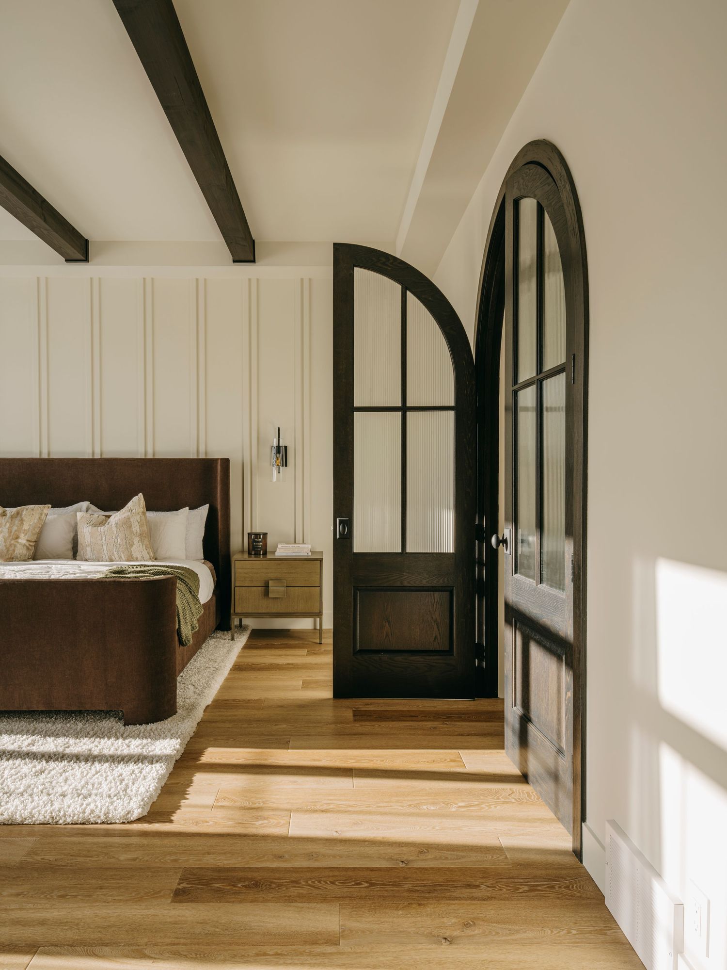 Elegant bedroom with arched black steel doors, wood beam ceiling, hardwood floors, and dark wood bed frame.