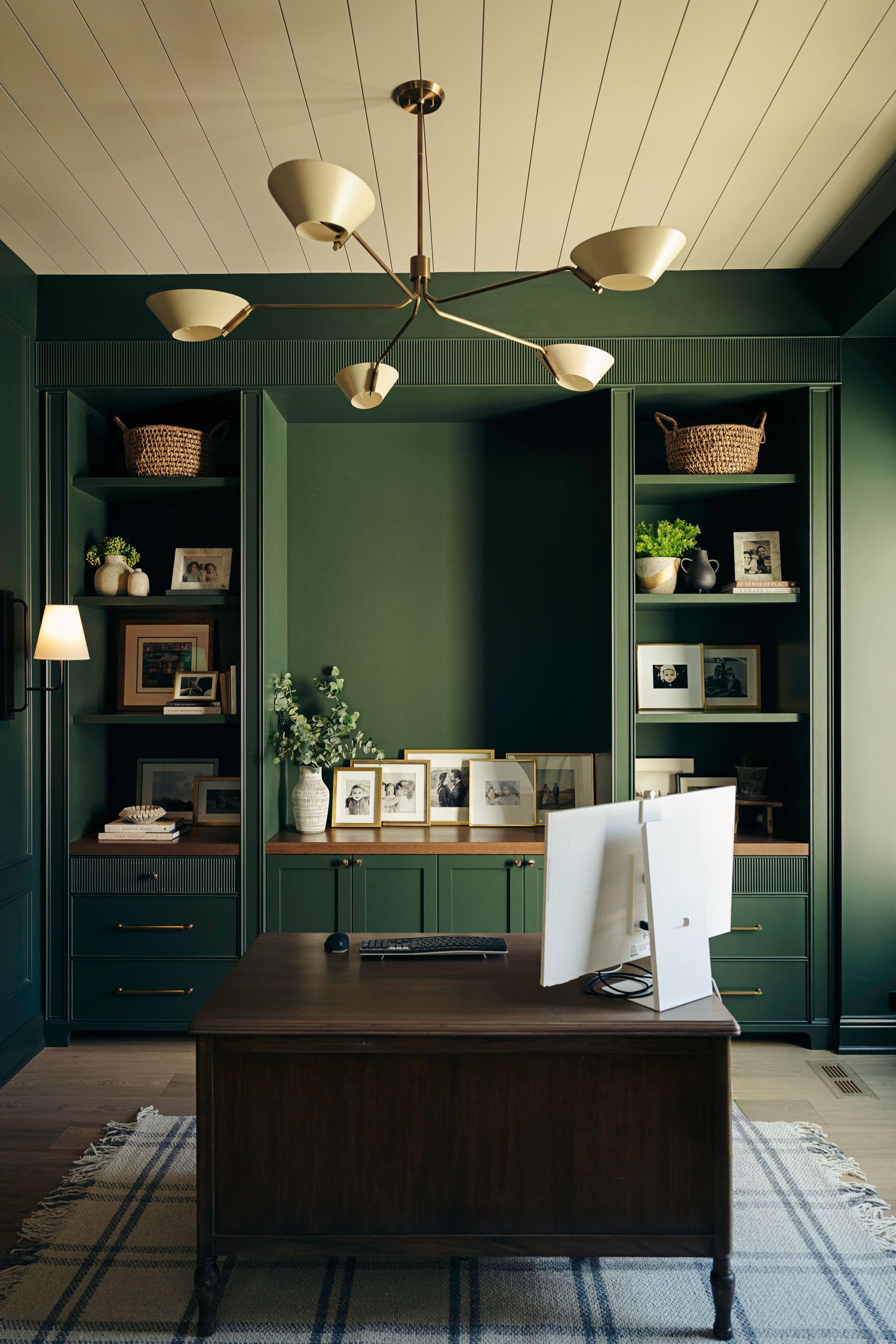 Dark green home office with built-in shelves, wood desk, brass chandelier, and decorative accents for an elegant workspace.