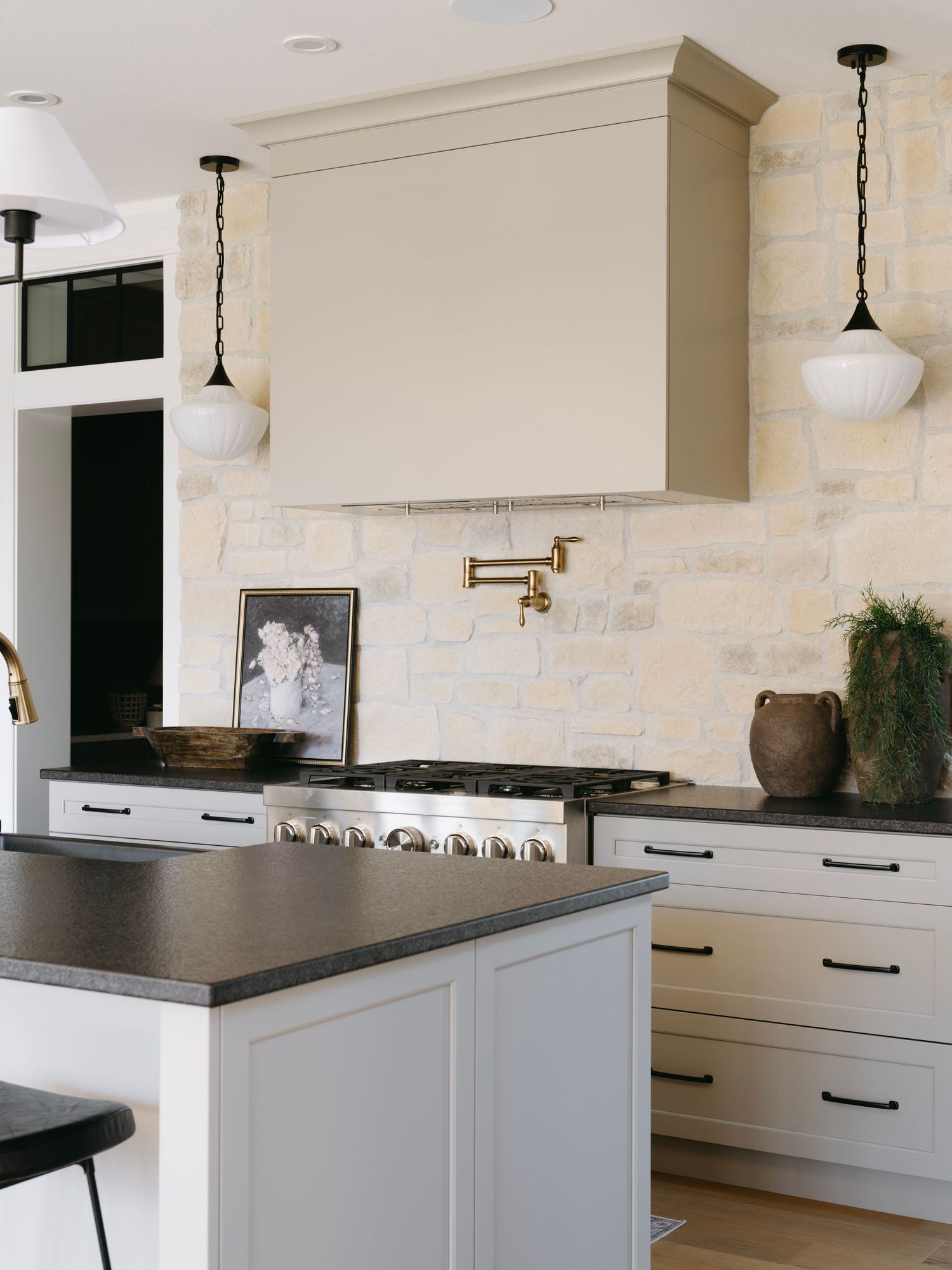 Classic kitchen featuring cream range hood, stone tile backsplash, dark countertops, brass pot filler, and globe pendant lights.