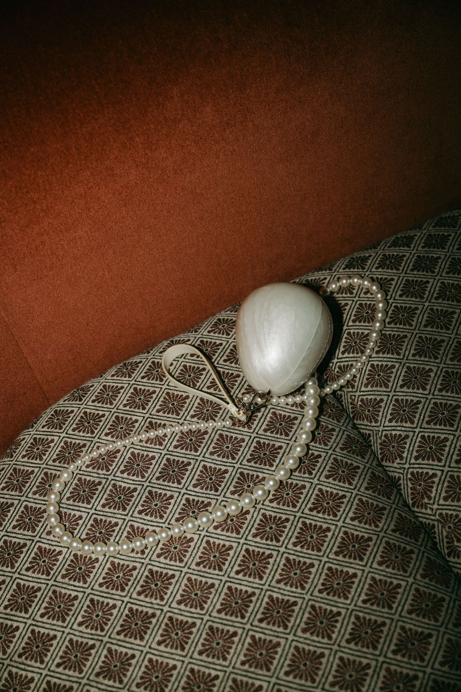 Pearl necklace and white compact mirror resting on vintage patterned cushion with terracotta velvet background.