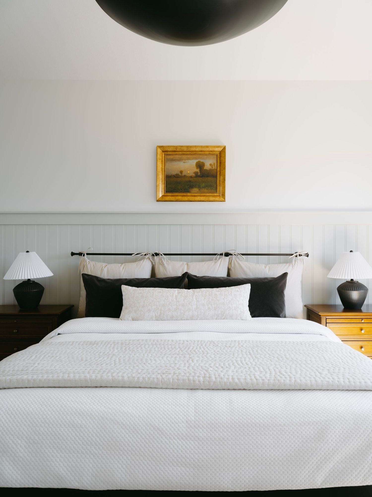 Classic white bedroom with paneled headboard wall, brass rail, landscape painting, black lamps, and dark sphere pendant.