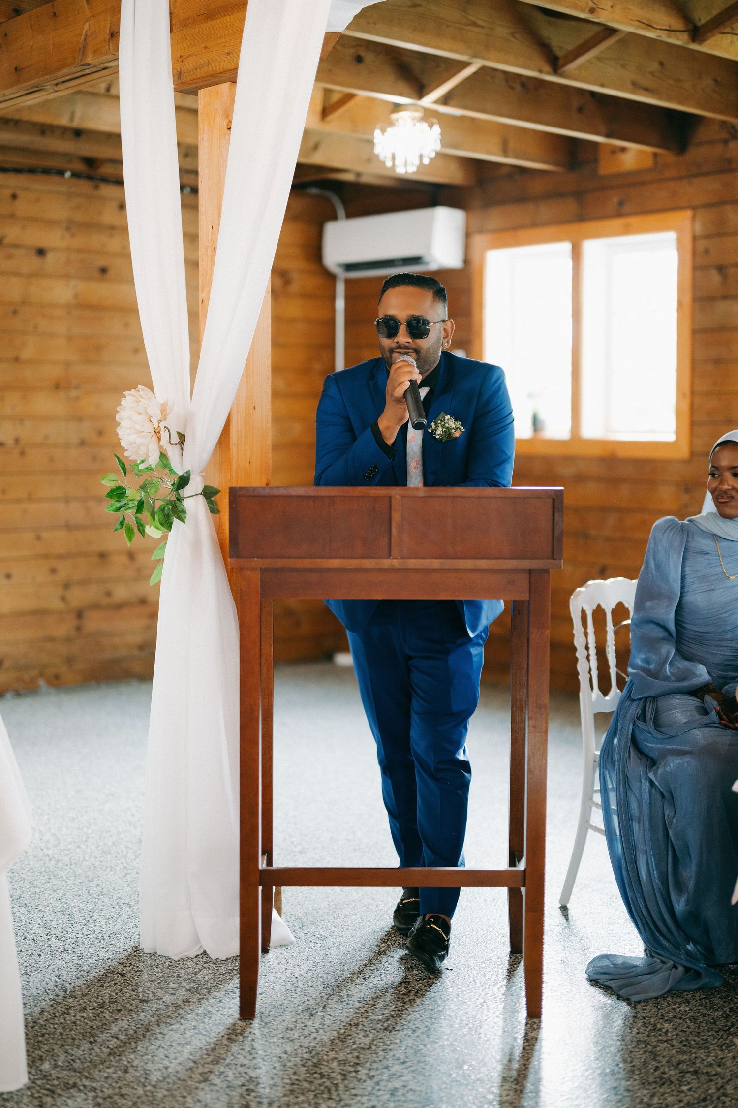Groom in blue suit speaking at wooden podium during indoor wedding ceremony.