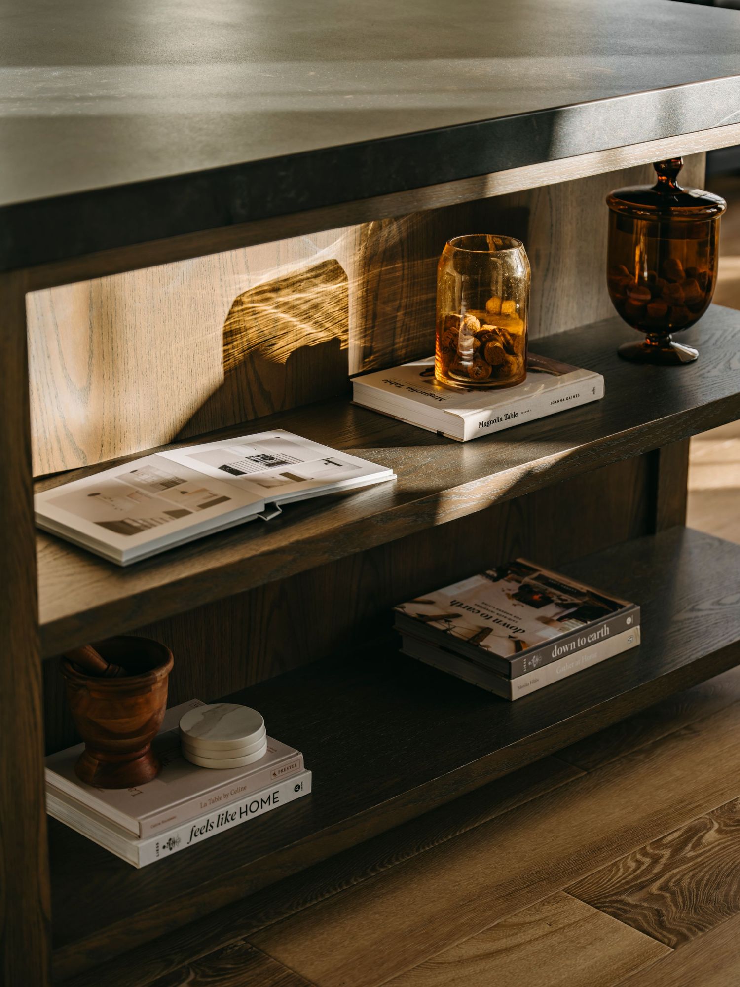 Dark wood bookshelf styled with open books, amber glassware, wooden bowl, and candle in warm natural light.