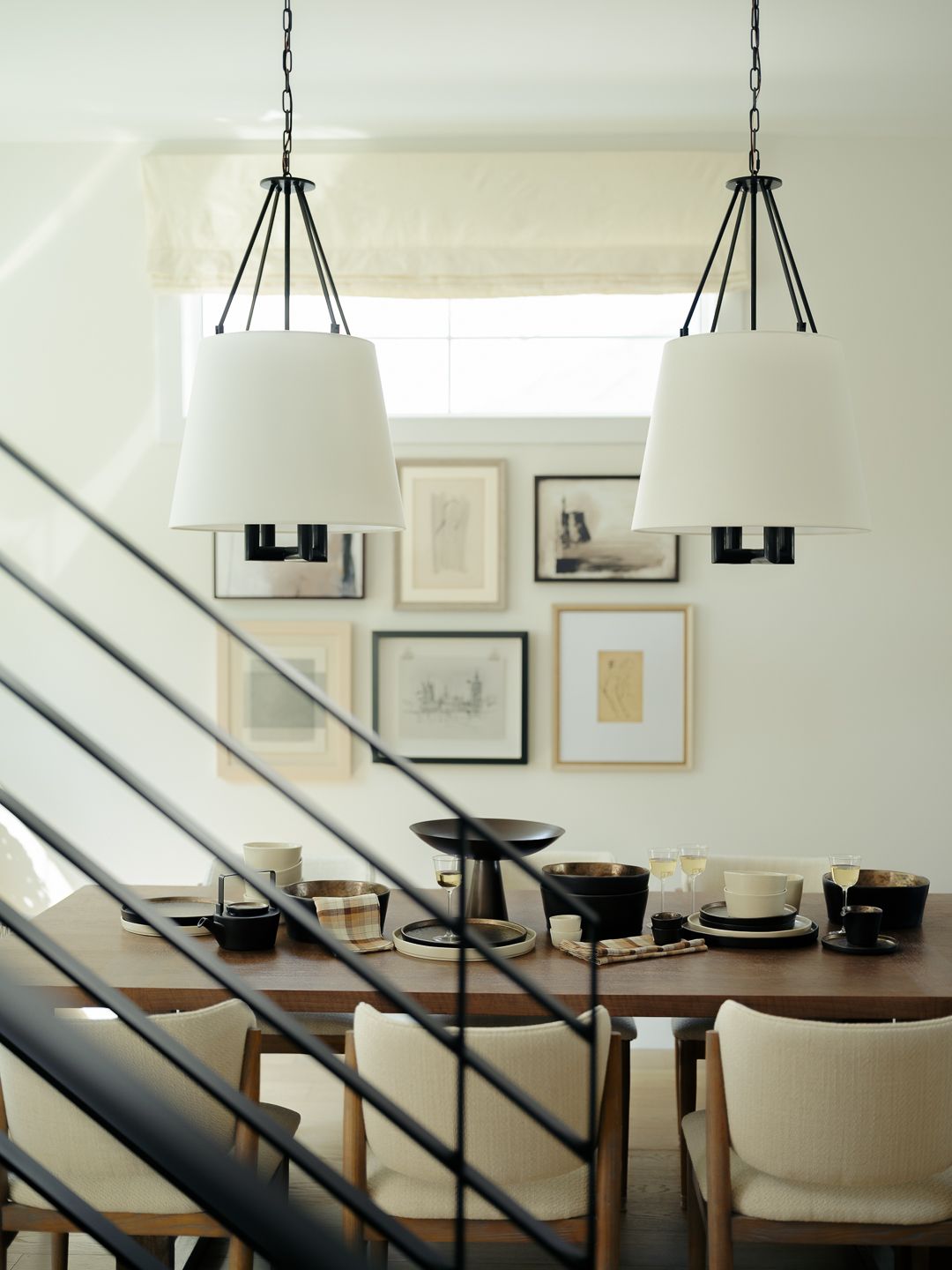 Elegant dining room with black pendant lights, white shades, wood table, upholstered chairs, and gallery wall art near a staircase.