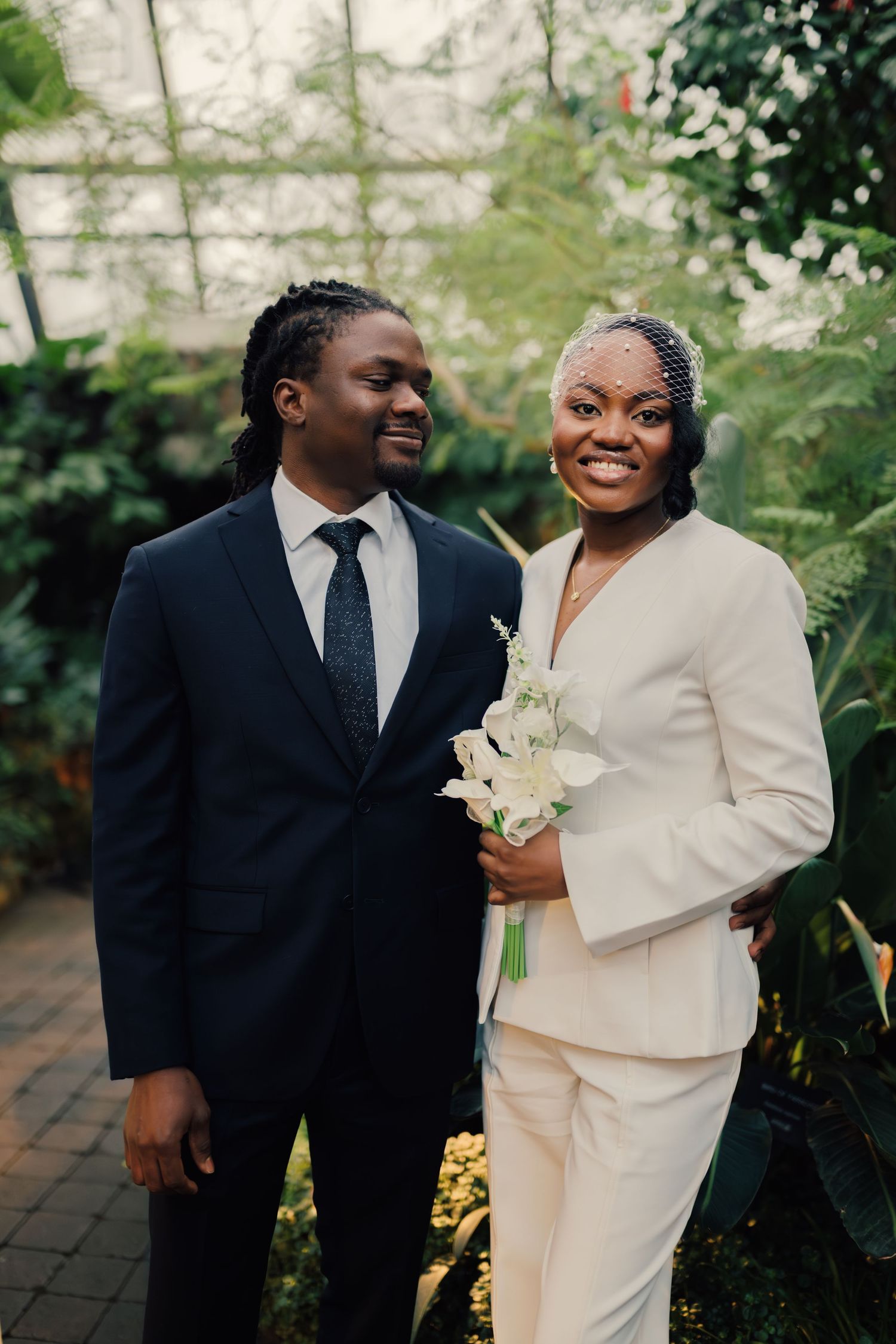 Wedding couple sharing a joyful moment in tropical greenhouse, bride in white suit with elegant bouquet.