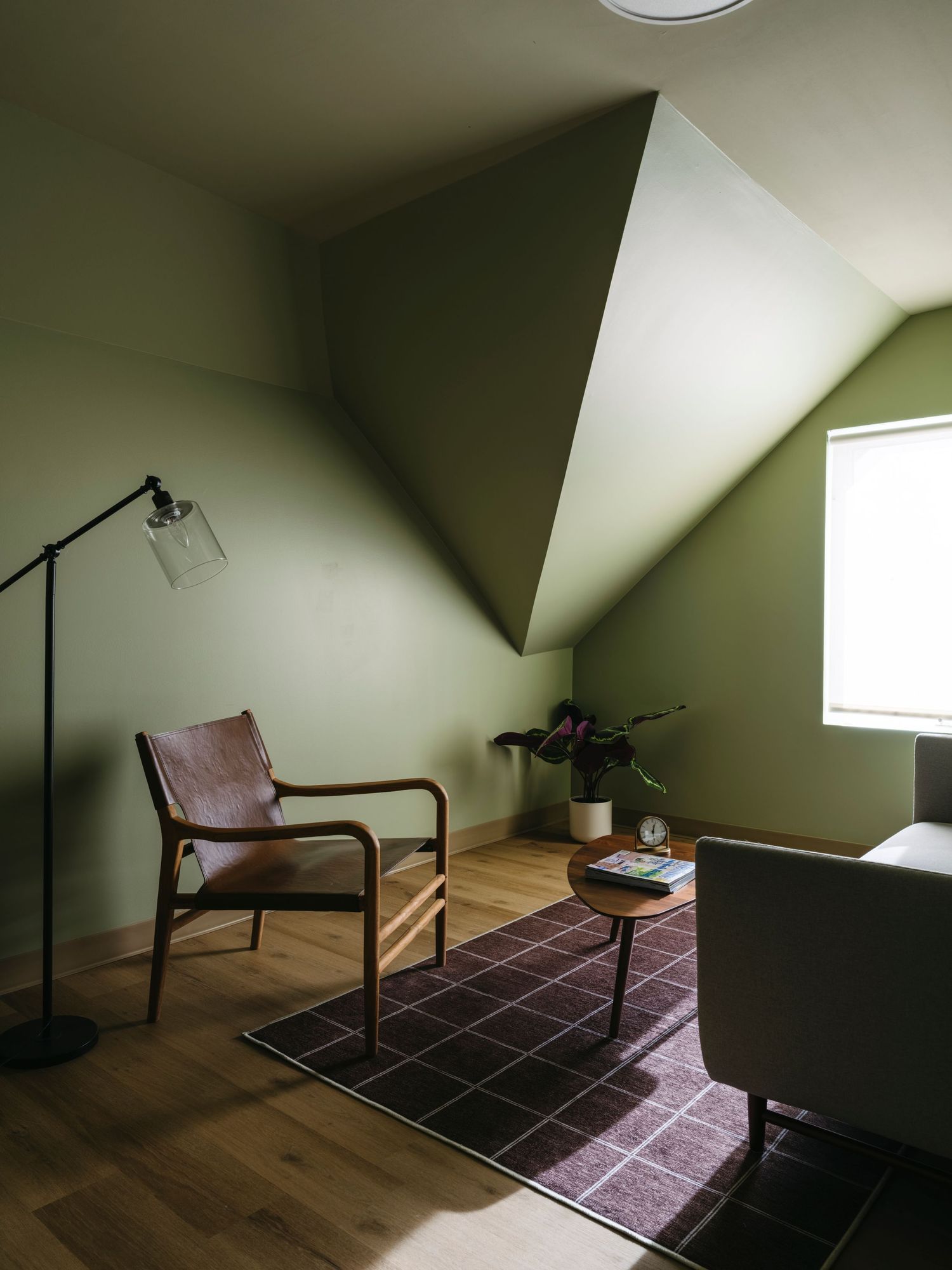 Cozy attic sitting room with olive green walls, leather chair, floor lamp, and plaid area rug.