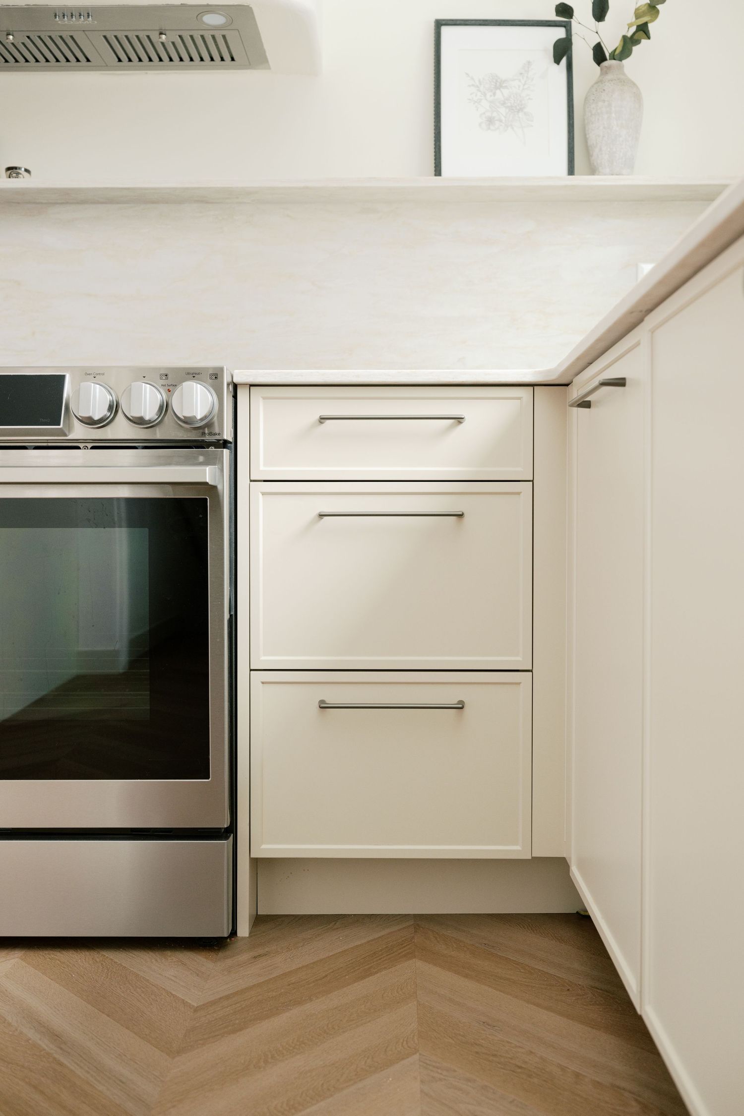 Modern white kitchen corner with stacked drawers, marble countertop, herringbone wood floor, and stainless steel oven.