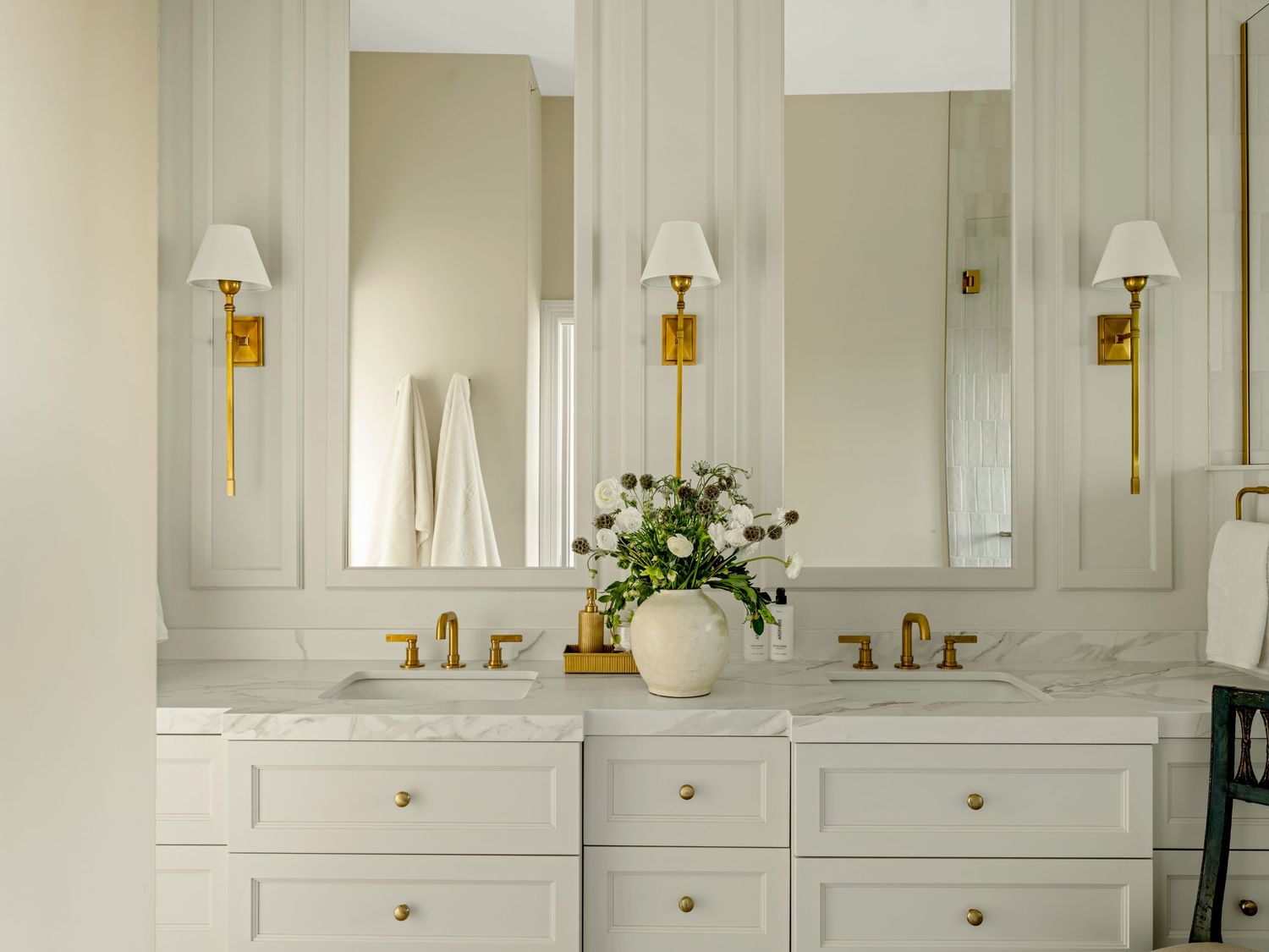 Elegant white double vanity bathroom with gold fixtures, marble countertops, wall sconces, and framed mirrors.