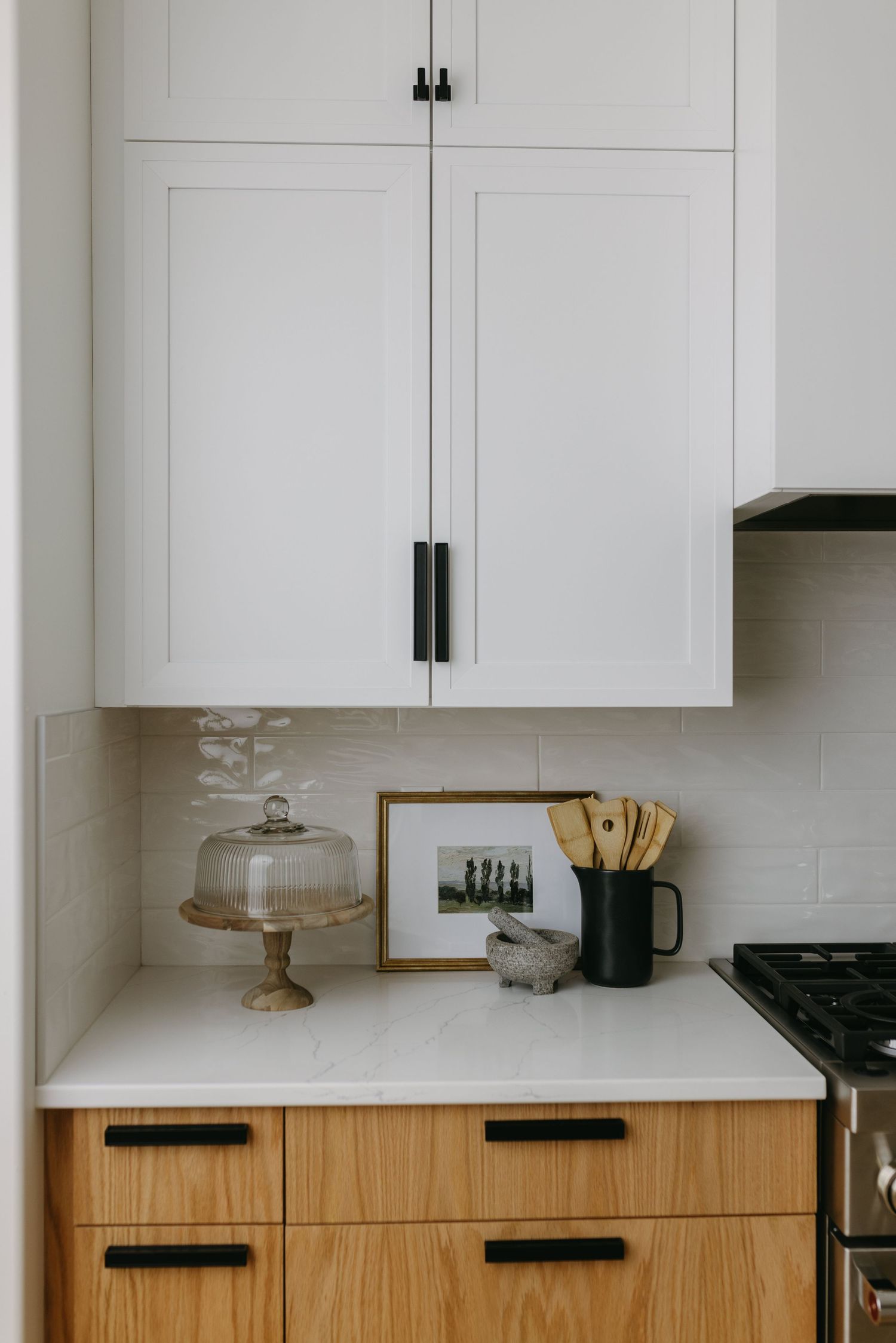 Modern kitchen with white shaker cabinets, wood lower drawers, black hardware, marble countertop, and styled decor including a cake stand.