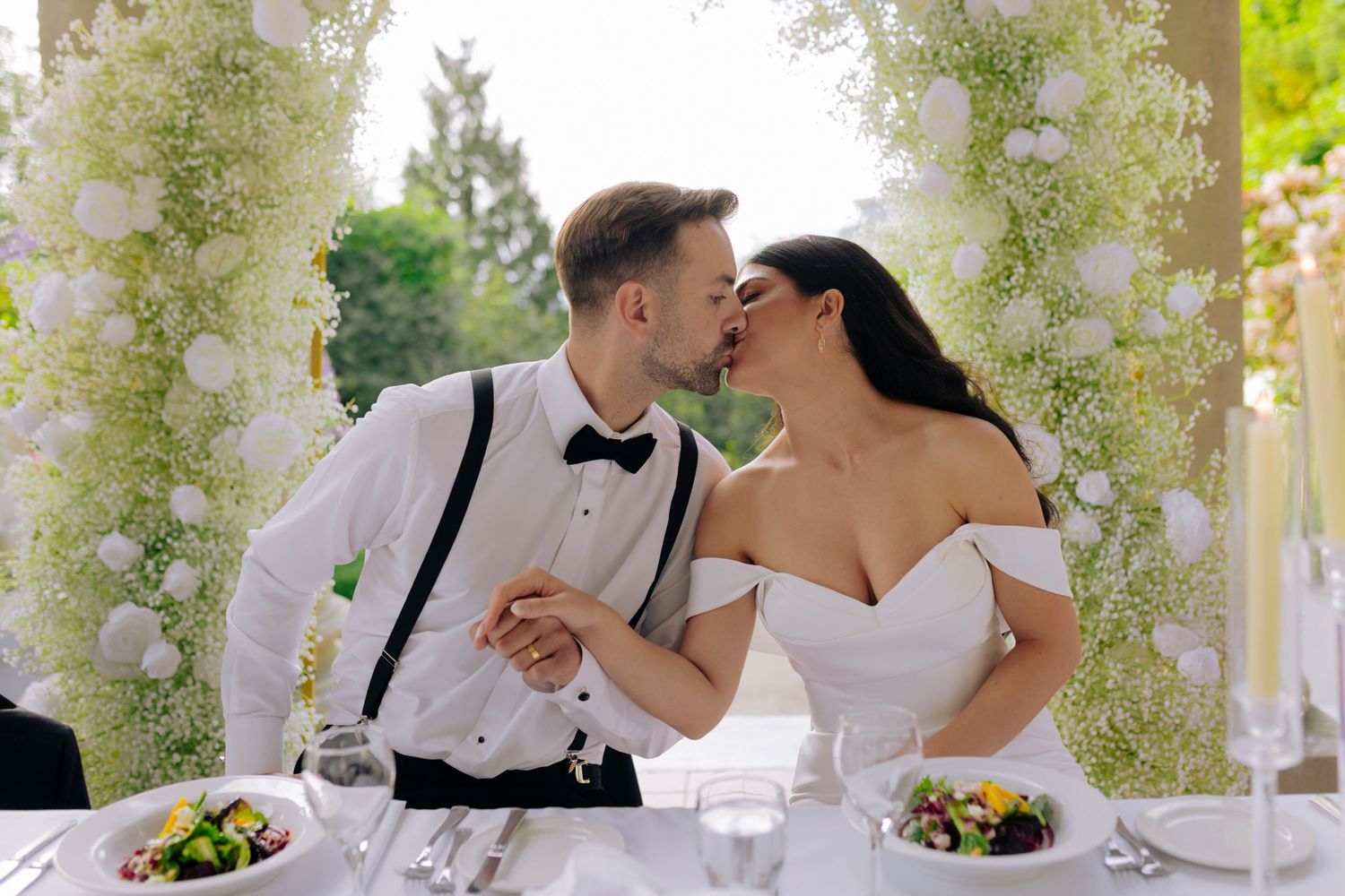 Bride and groom sharing a kiss at outdoor reception table surrounded by white floral arrangements.