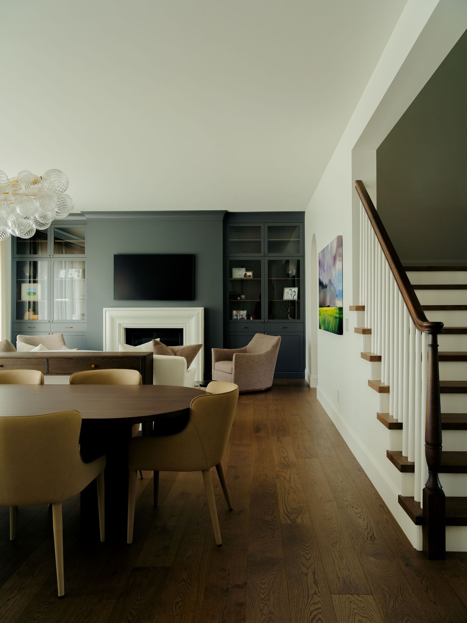 Modern open-plan living and dining room with dark walls, fireplace, built-in shelving, hardwood floors, and elegant staircase.