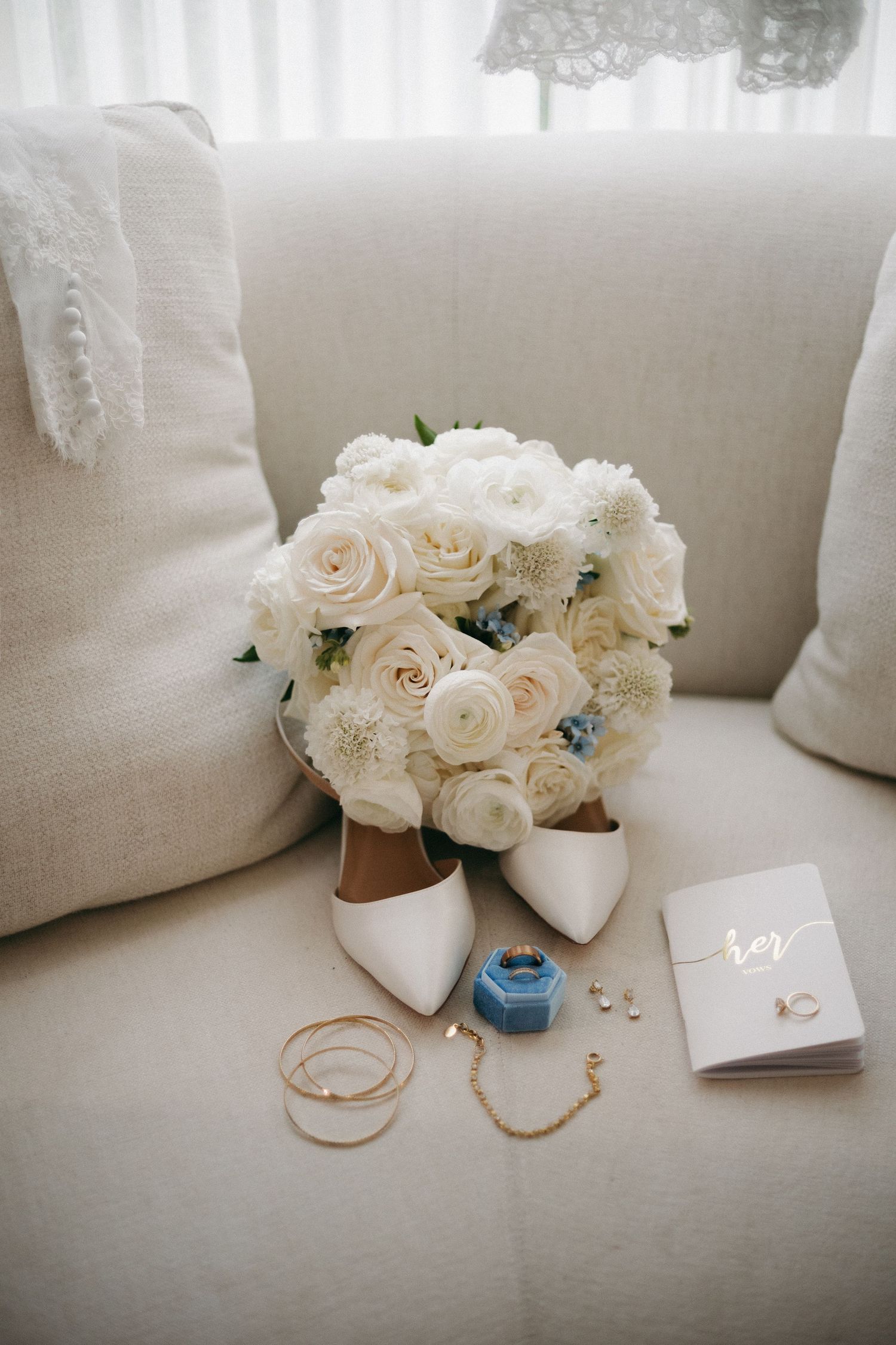 White rose bridal bouquet with wedding shoes, blue ring box, gold jewelry, and vow booklet on cream sofa.